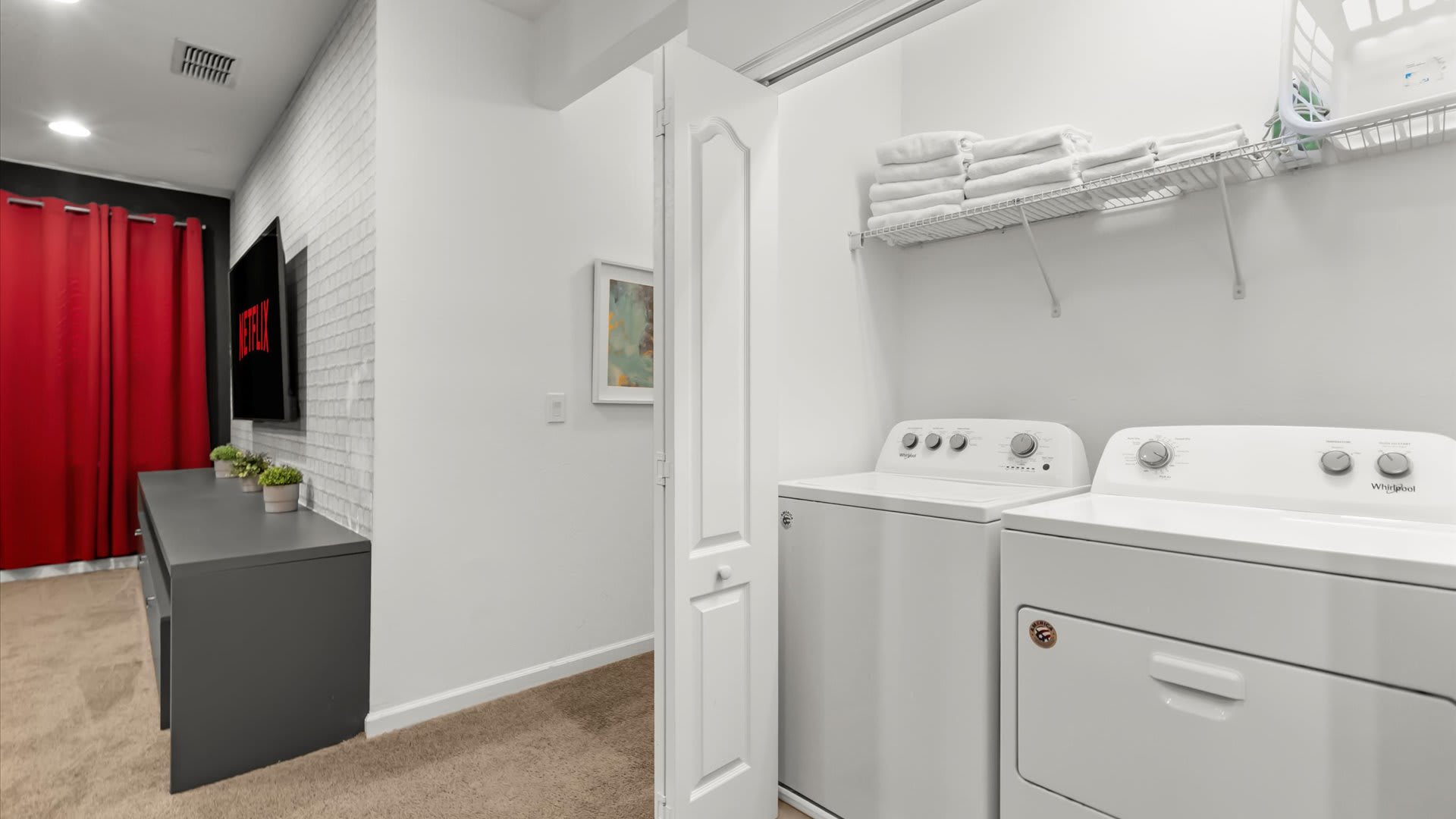 Bright laundry closet with white Whirlpool washer and dryer, a wire shelf holding neatly folded white towels, and a partial view into an adjacent media room with a dark entertainment console and a TV displaying Netflix.