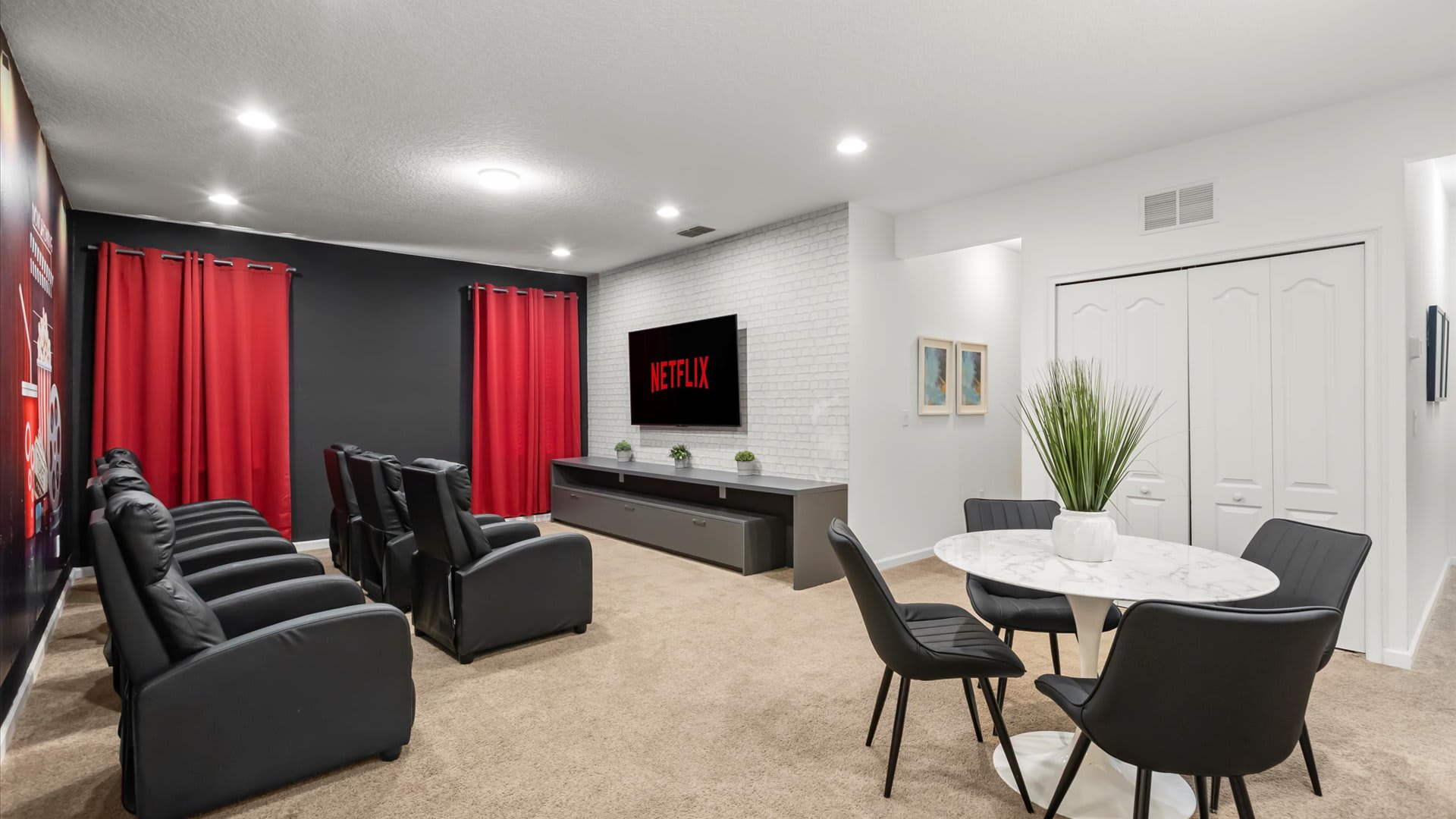 A dedicated media room features multiple rows of black leather recliners facing a large wall-mounted TV displaying "NETFLIX." The room has dark gray walls, red curtains, and a white brick accent wall, with a small white dining table and black chairs in the foreground.