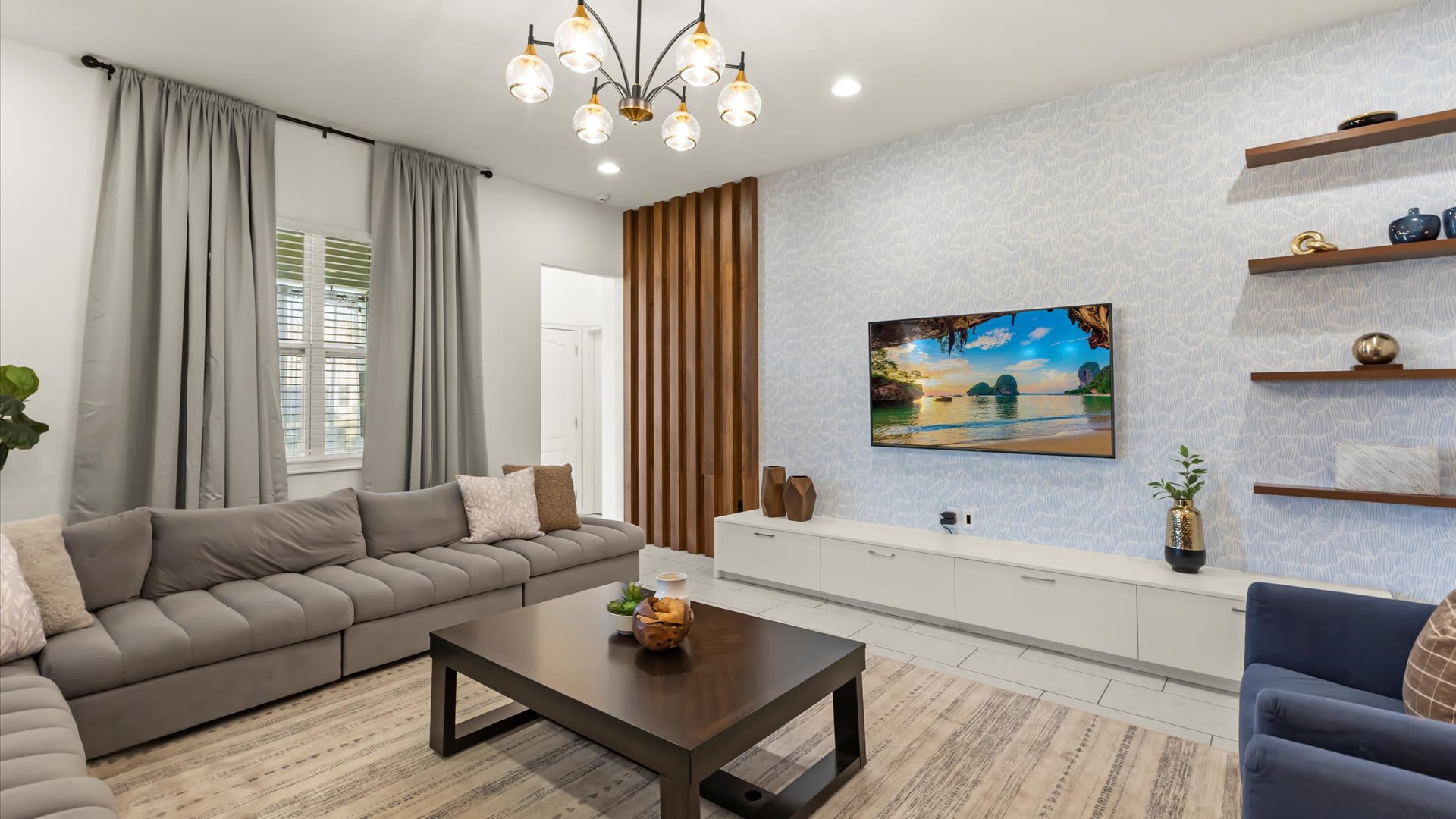 A contemporary living room features a large gray sectional sofa and a dark wood coffee table on a striped rug. A wall-mounted TV is set against a light blue patterned accent wall, flanked by floating shelves and a decorative wooden slat partition.