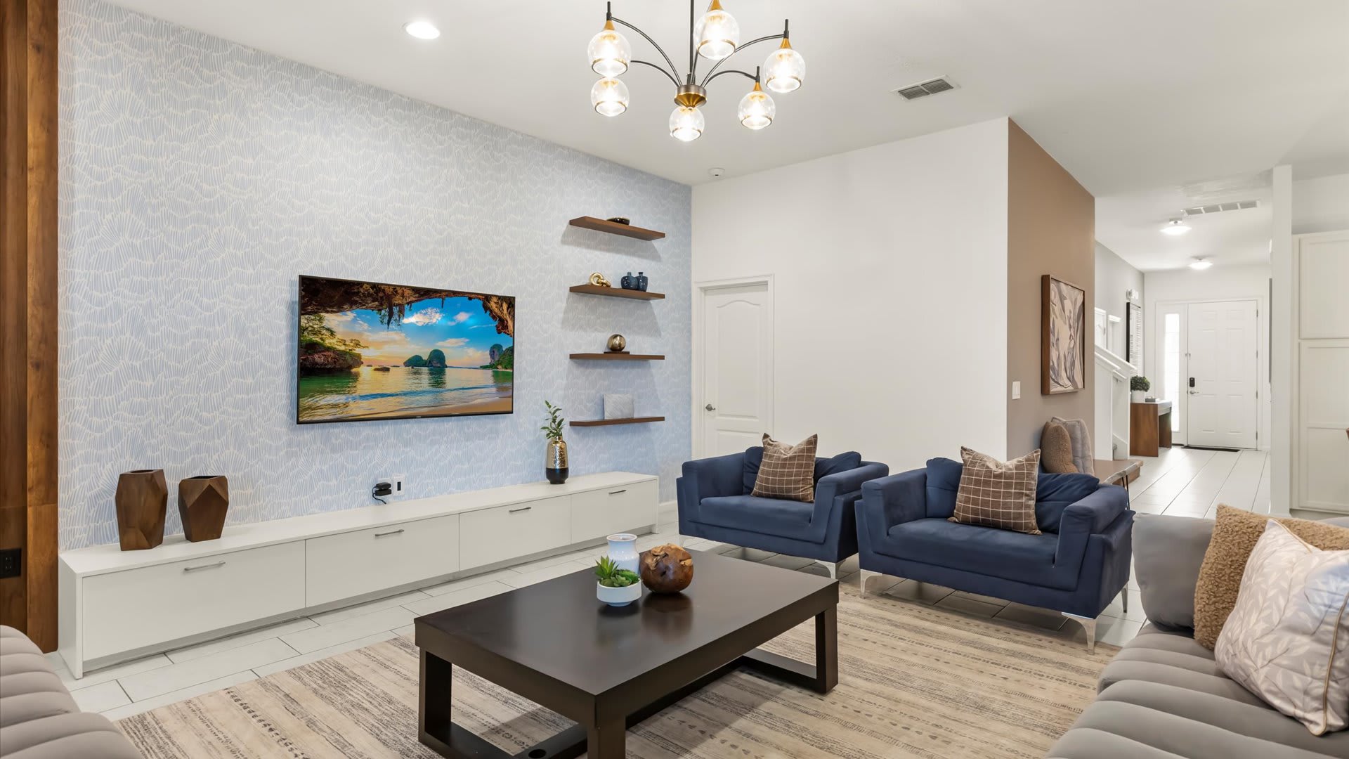 This bright living room showcases a gray sectional sofa, two blue armchairs, and a dark wood coffee table on a patterned rug. A large TV is mounted on a light blue patterned accent wall, complemented by floating shelves and a modern chandelier.