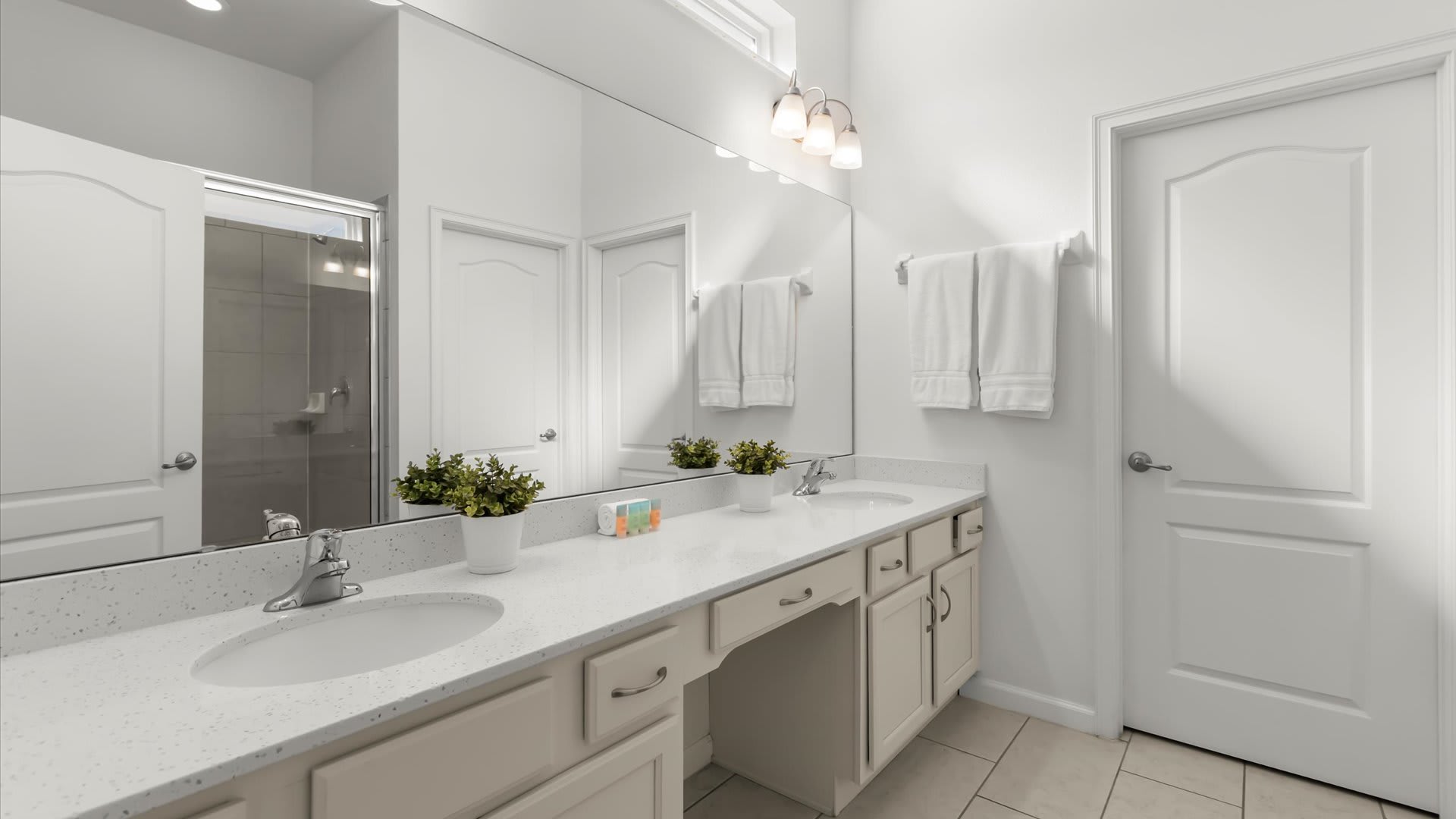 A bright, modern bathroom featuring a double vanity with white quartz countertops, two sinks, and a large mirror reflecting a glass-enclosed shower. The room has white walls, light floor tiles, and contemporary vanity lighting.