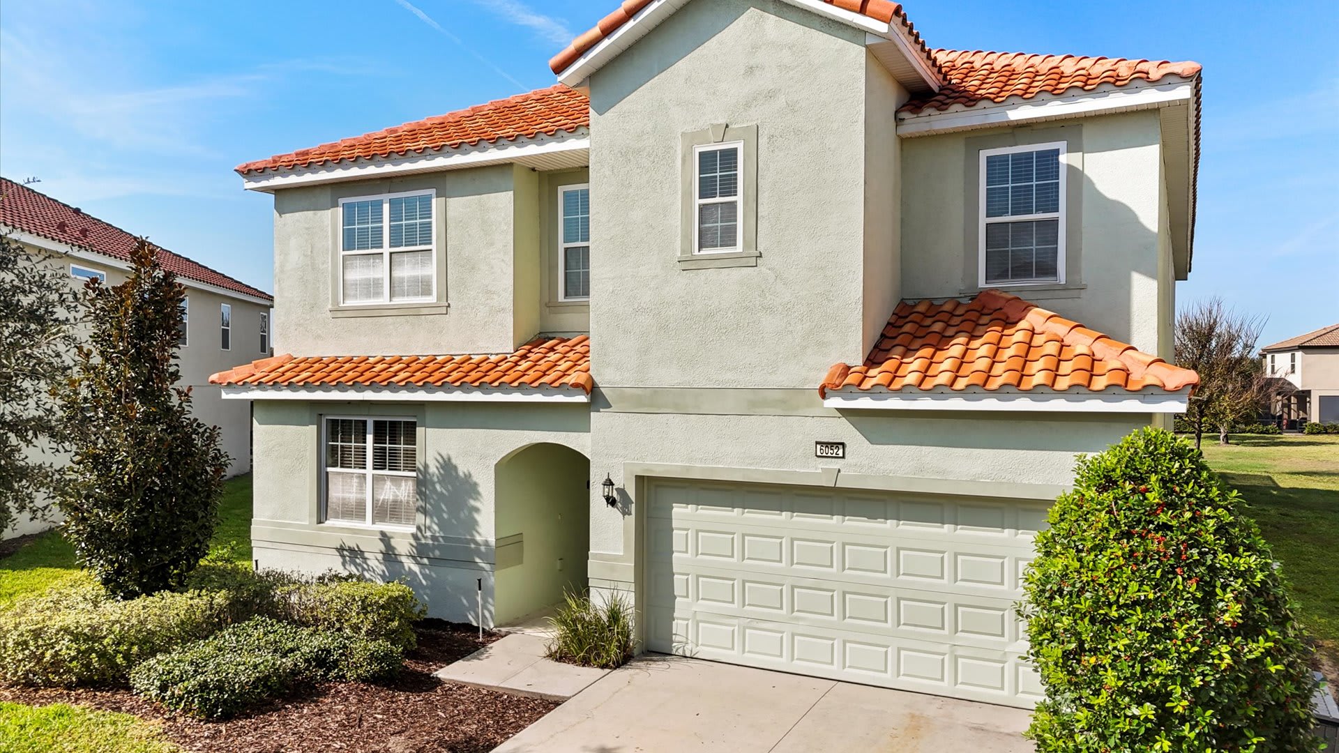 Two-story Mediterranean-style house with light sage green stucco exterior, a distinctive terracotta tile roof, multiple white-framed windows, and an attached garage, fronted by a paved driveway and well-maintained landscaping.