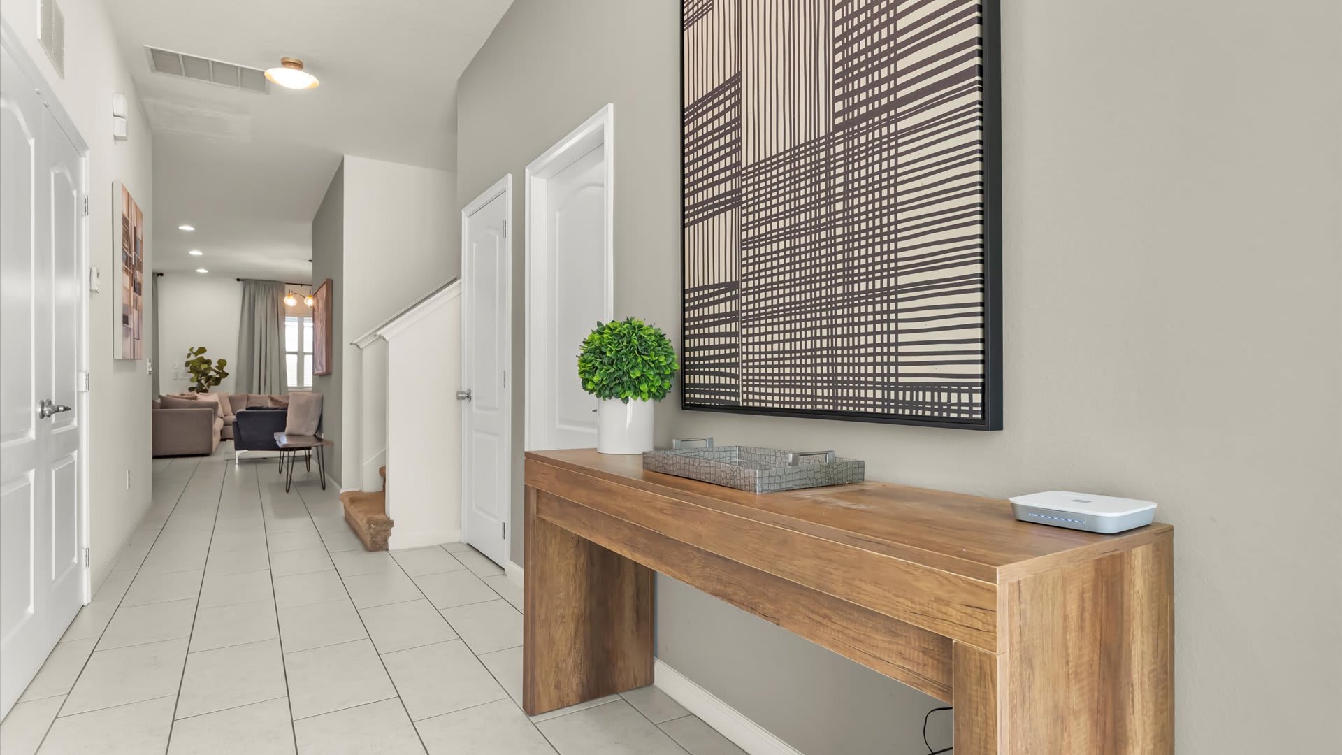 Bright and modern foyer featuring light gray walls, white tiled flooring, and a rustic wooden console table adorned with a green potted plant and a large abstract black and white artwork. The hallway leads to a living area with a glimpse of a staircase.