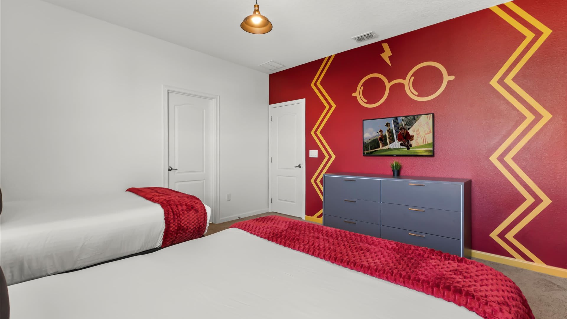 Another view of the Harry Potter-themed bedroom, showcasing a red accent wall with a lightning bolt and glasses design, a wall-mounted flat-screen TV, and two queen beds with red throws.