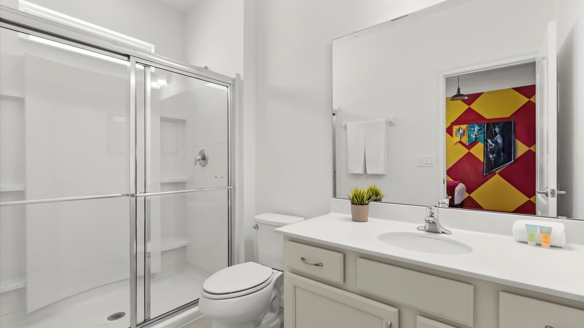 Modern white bathroom featuring a single vanity with a large mirror, a toilet, and a spacious glass-enclosed shower with a built-in bench. The mirror reflects a brightly themed room with yellow and red checkered walls.