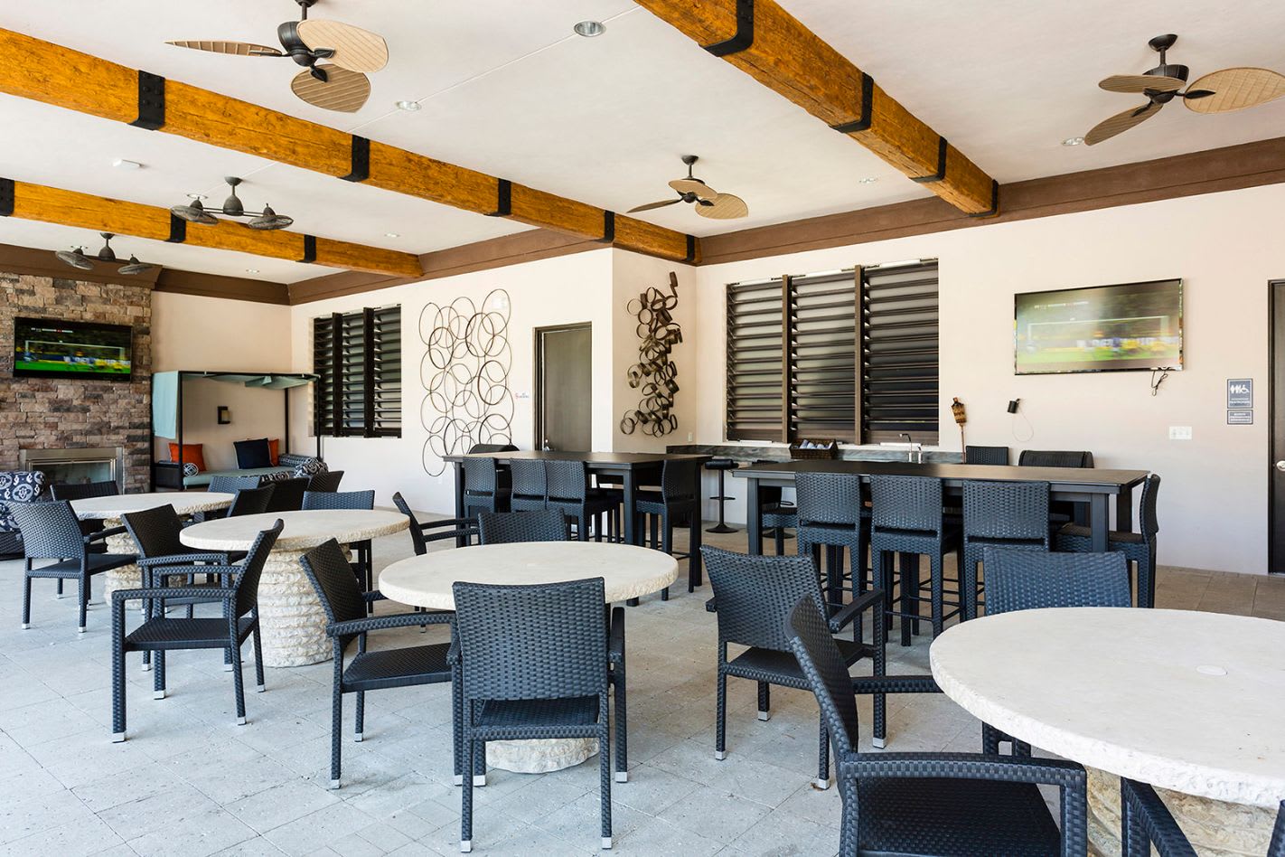 Spacious and airy outdoor covered lounge area, featuring multiple seating arrangements with round and rectangular tables and black wicker chairs, exposed wooden beam ceiling with ceiling fans, and wall-mounted televisions.