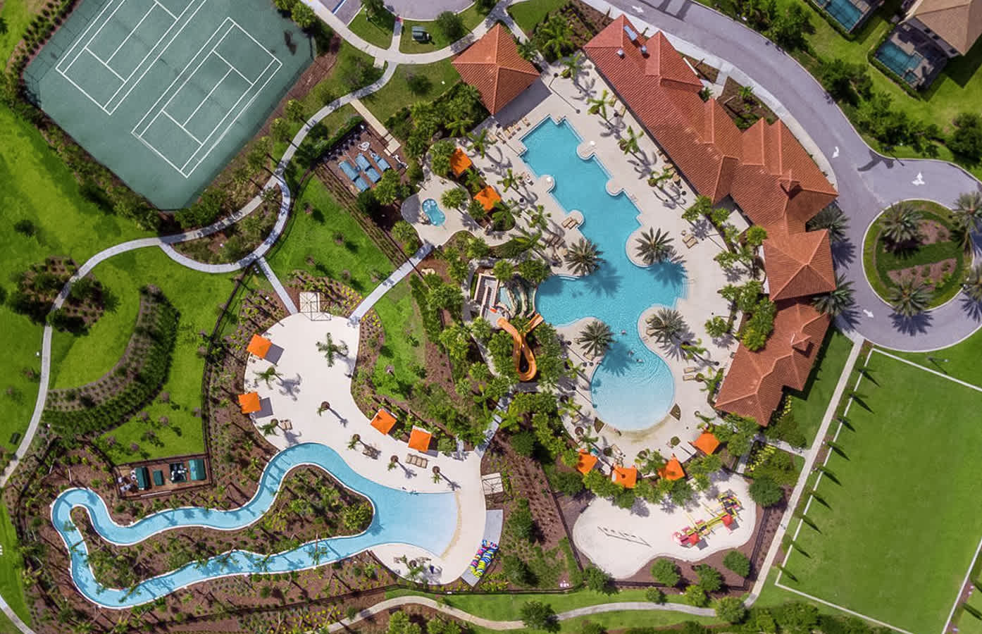 Comprehensive aerial shot of a vibrant resort amenity area, highlighting a large communal pool with water slides, a lazy river, tennis courts, and lush green spaces, all interconnected by paved pathways.