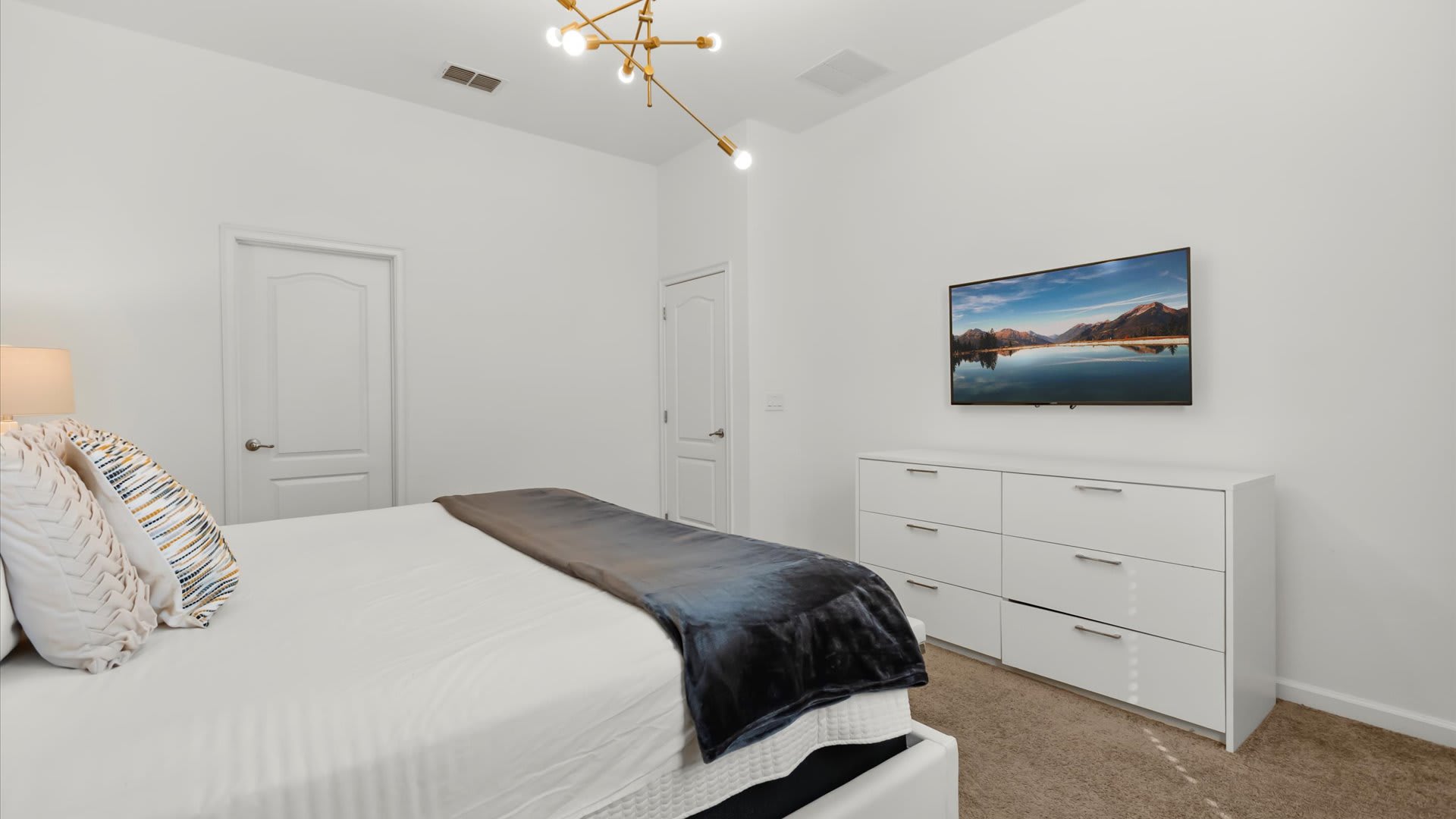 View of the foot of the bed in bedroom-3, showing a white dresser with a wall-mounted flat-screen TV displaying a mountain lake scene. The room has white walls and a light brown carpet.