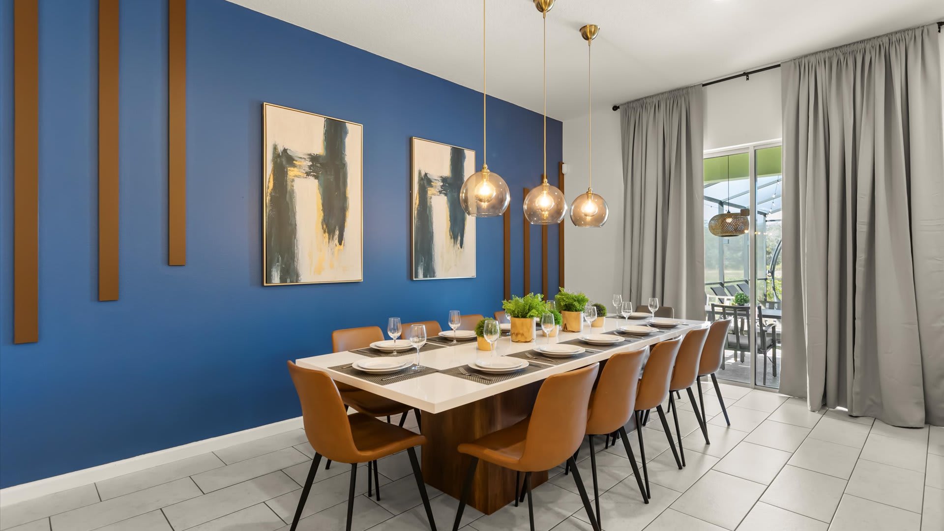 An elegant dining room features a long white dining table with seating for ten, adorned with modern place settings and greenery. The vibrant blue accent wall is decorated with two abstract art pieces and vertical wooden slats, illuminated by three contemporary pendant lights.