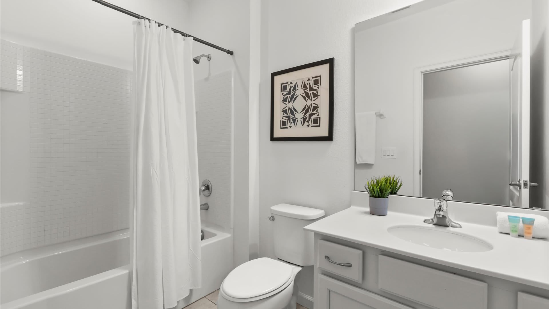 Bright white bathroom with a single vanity, toilet, and a shower-tub combination enclosed by a white curtain. A framed abstract artwork hangs above the toilet.