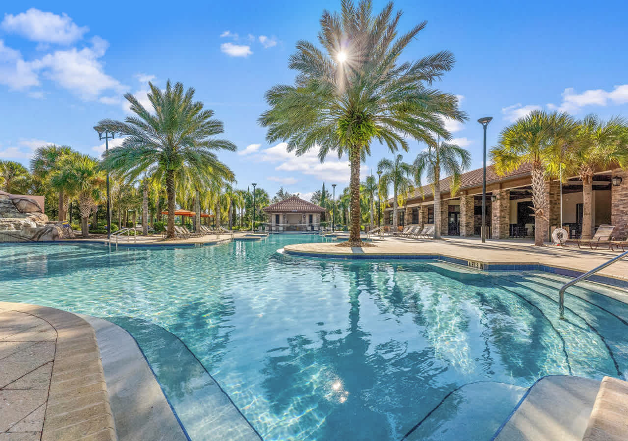 Picturesque communal pool scene with sparkling blue water, framed by tall, swaying palm trees and a paved deck with lounge chairs, bathed in bright sunlight under a partly cloudy sky.
