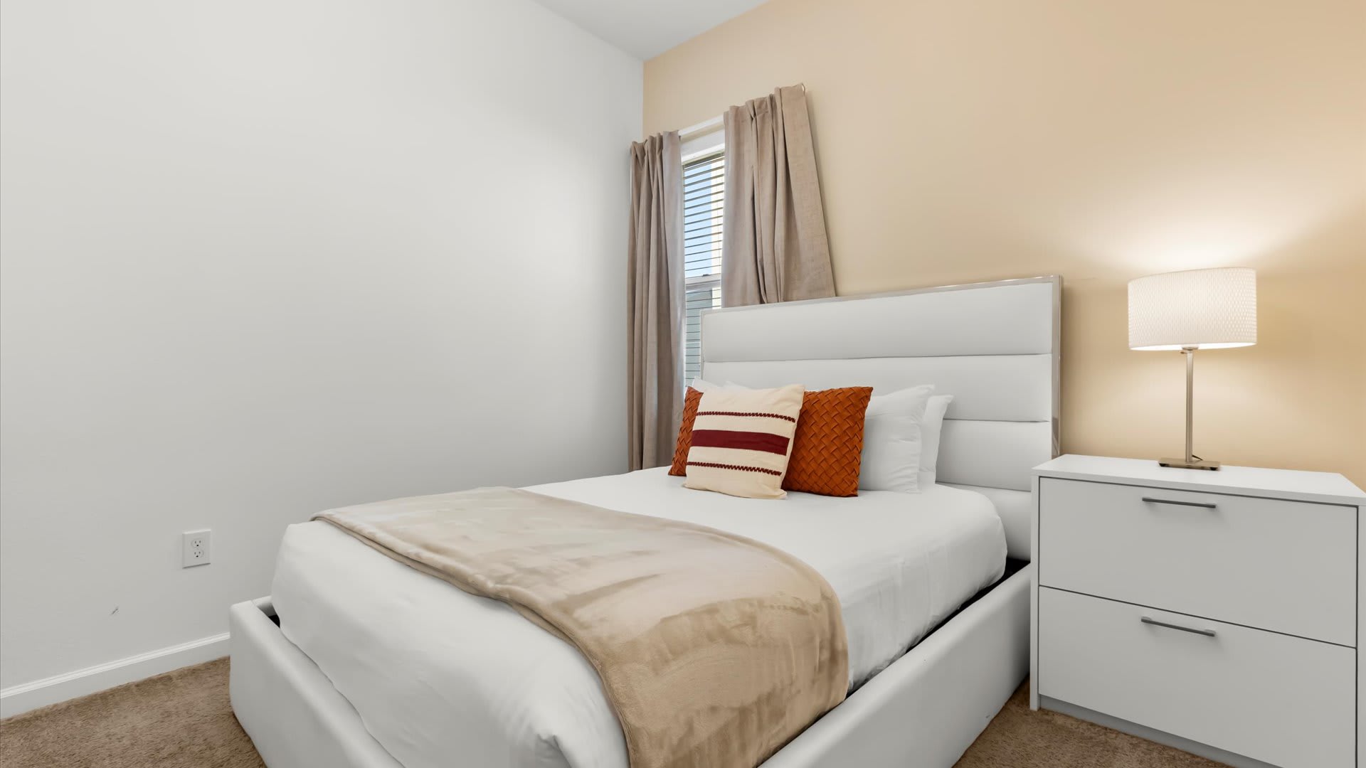 A bright bedroom with a white upholstered bed, a beige throw blanket, and orange and white decorative pillows, next to a white nightstand with a lamp. The room has a two-tone wall with white and light beige sections, and a window with beige curtains.