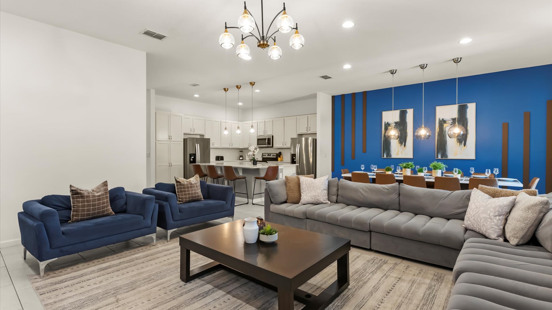 Bright and open living space featuring a large gray sectional sofa, a dark coffee table, and a striking blue accent wall, seamlessly connected to a modern kitchen with white cabinetry and a large island.
