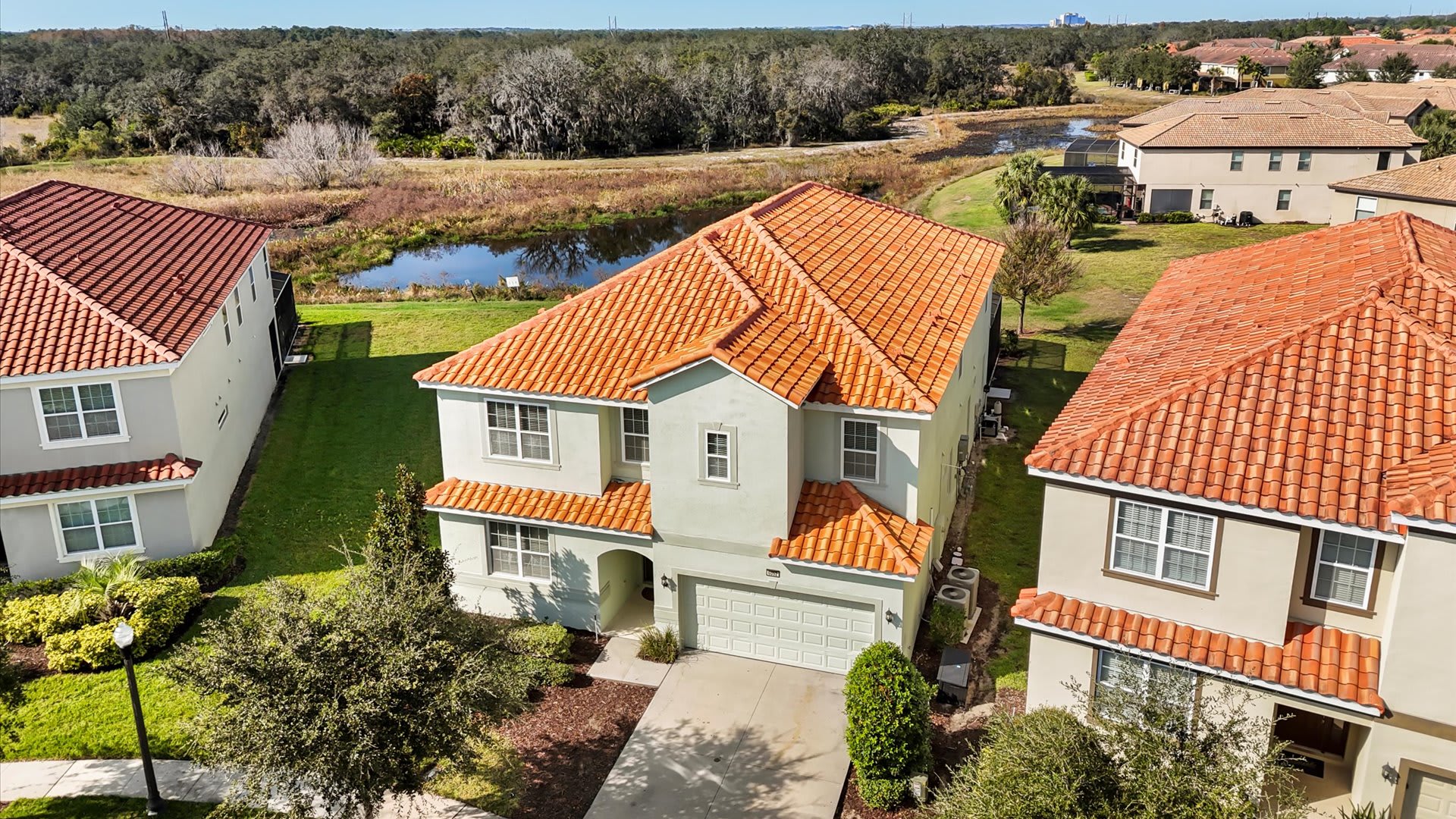 Aerial perspective of a two-story Mediterranean-style house with a terracotta tile roof, situated in a residential community, backed by a serene pond and natural wooded areas under a clear blue sky.