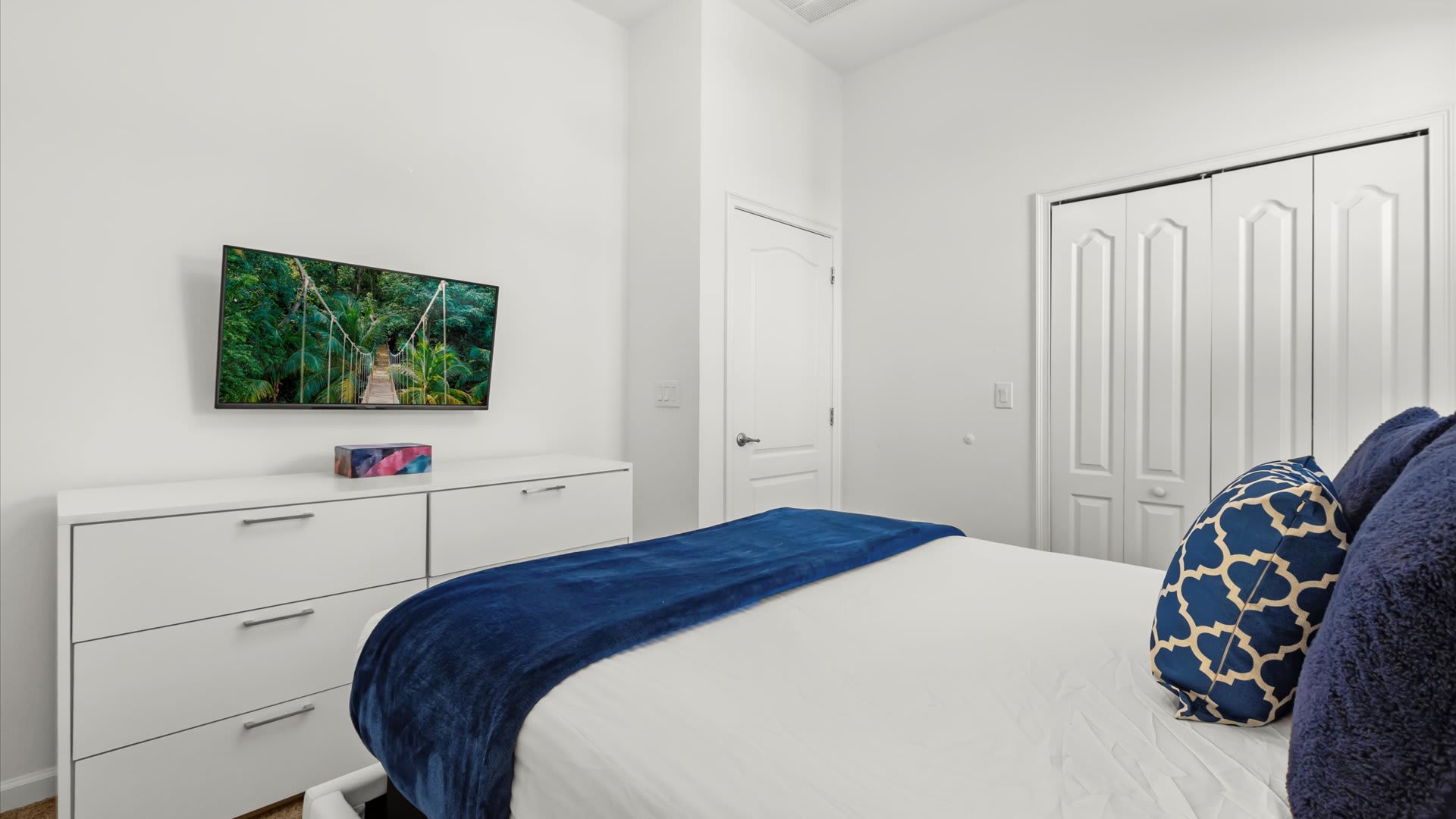 A modern bedroom wall featuring a flat-screen TV mounted above a white dresser with multiple drawers, positioned opposite a bed with a blue throw blanket. A white door and closet are visible.