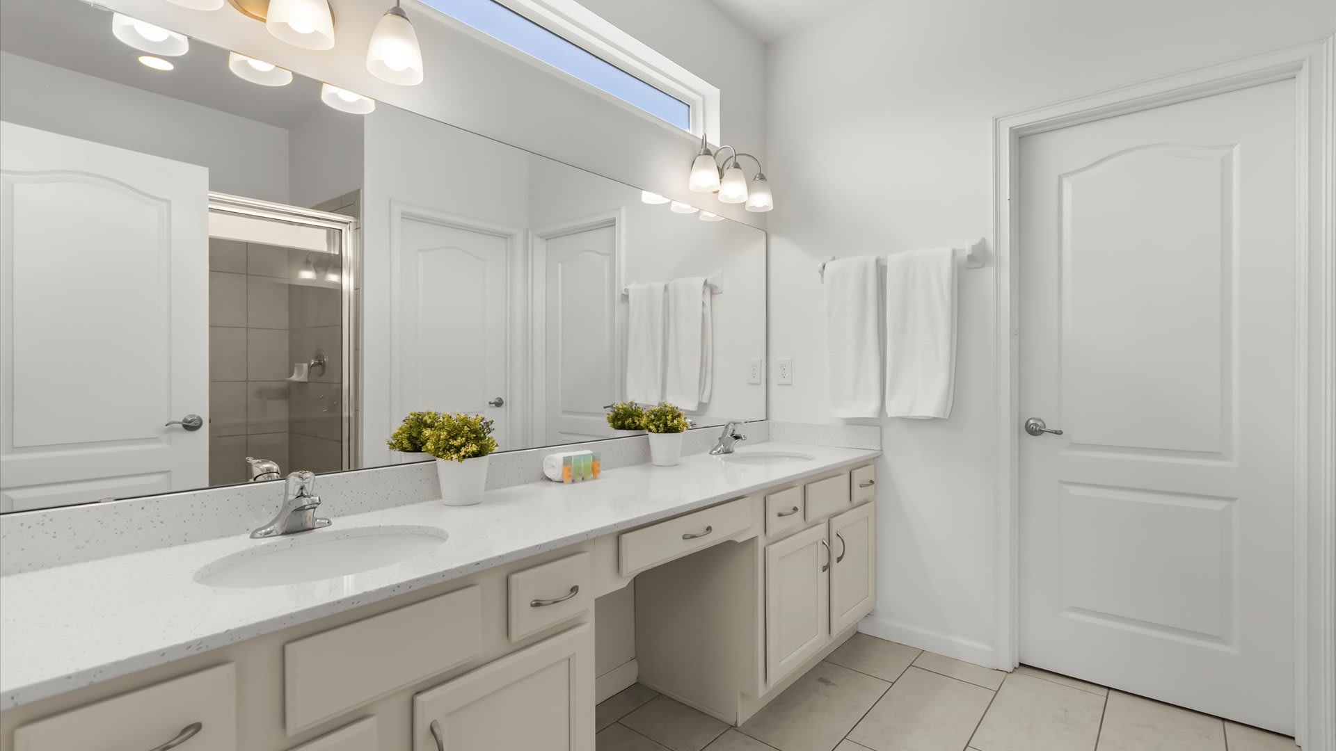 Modern bathroom featuring a long double vanity with white quartz countertops, two sinks, and light cream cabinets. A large mirror spans the length of the vanity, reflecting a glass-enclosed shower and white doors.