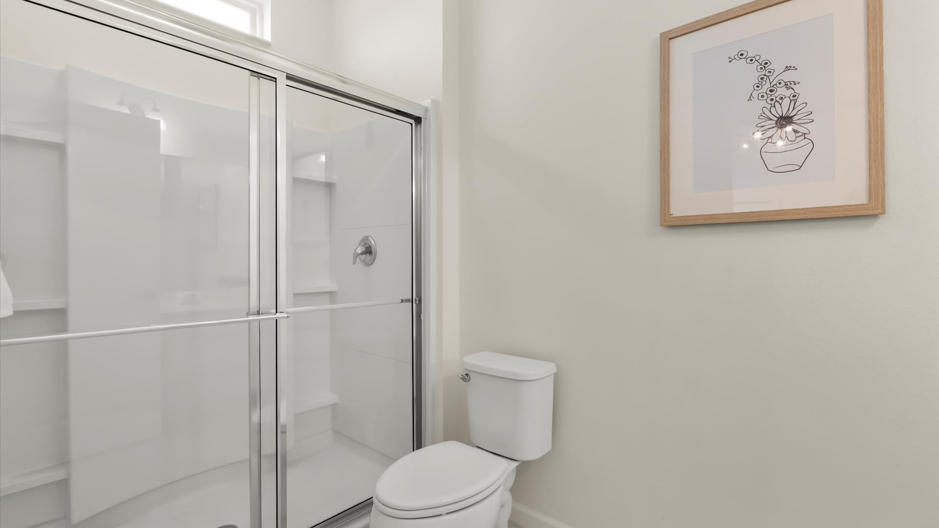 A clean, contemporary bathroom with a white toilet and a glass-enclosed walk-in shower featuring white subway tile walls. A framed abstract floral print hangs on the light beige wall.