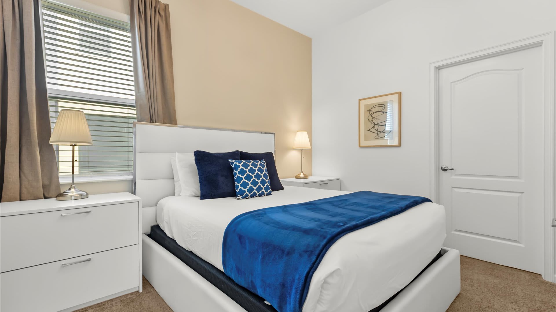 A bright bedroom featuring a white upholstered queen bed with a blue throw blanket and patterned pillows, flanked by two white nightstands with lamps. The room has a two-tone wall with white and light beige sections, and a window with beige curtains.