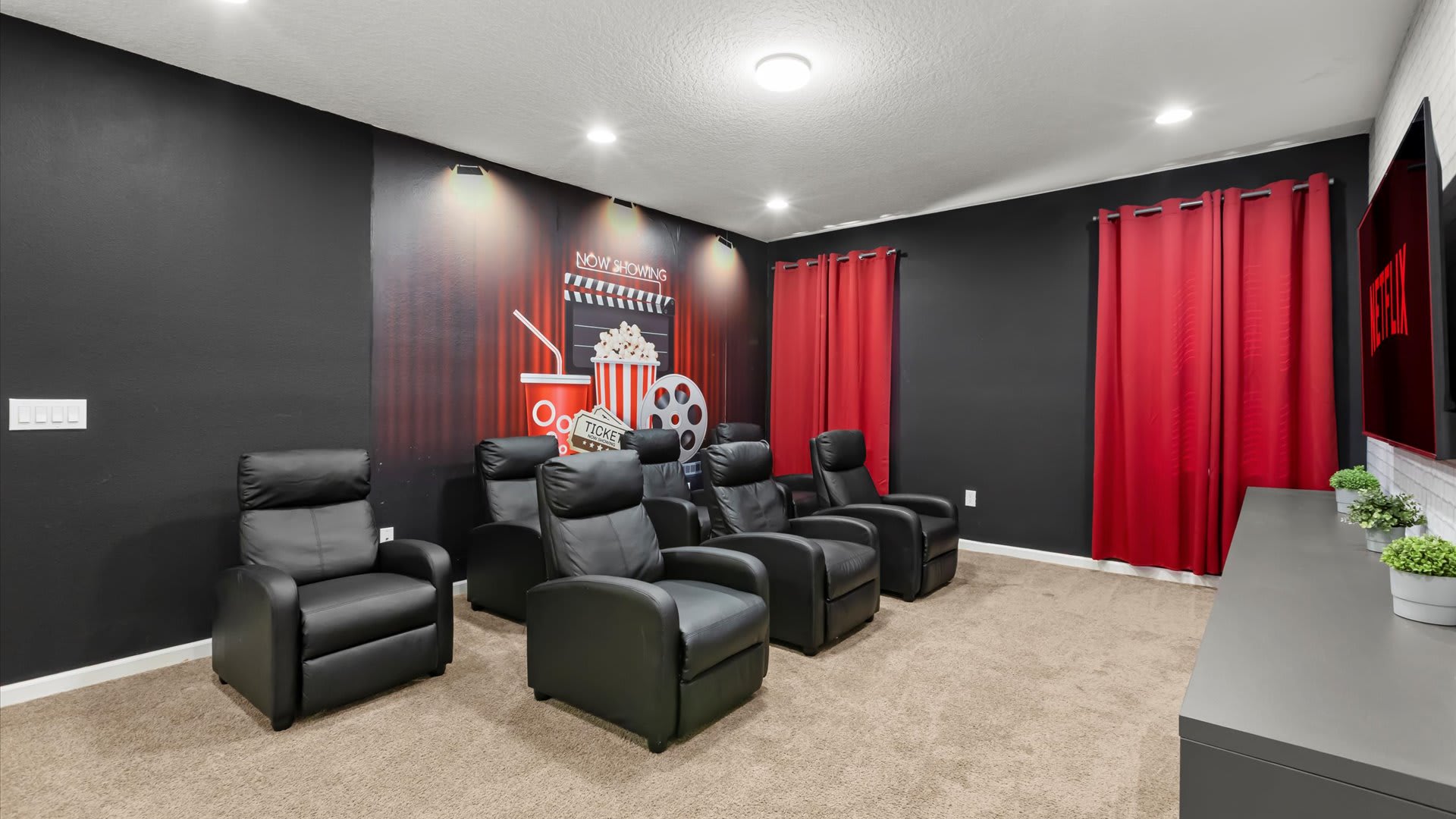 Dedicated home theater room with plush black reclining chairs arranged in rows, dark walls, red curtains, and a large movie-themed mural behind the seating area, creating an immersive viewing experience.