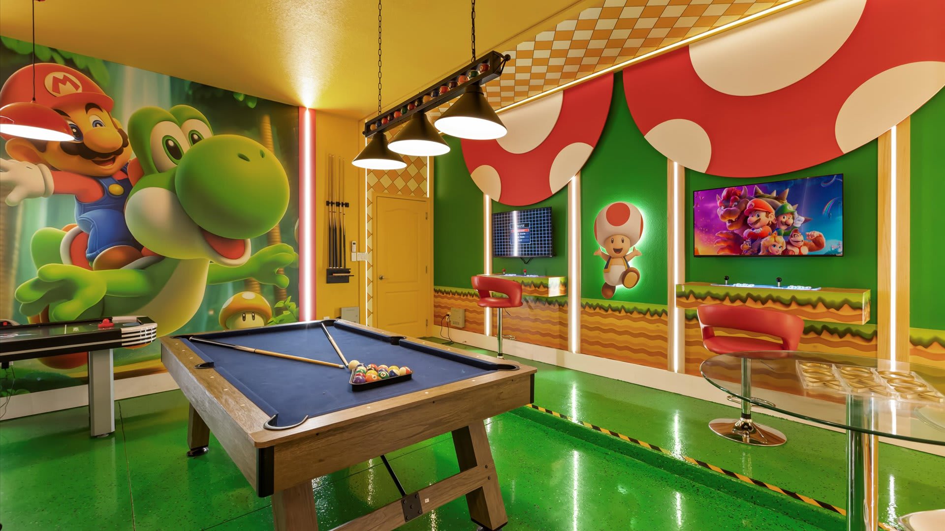 Vibrant Super Mario-themed game room featuring a pool table with blue felt, an air hockey table, and a foosball table, all set on a glossy green floor with character murals on the walls.