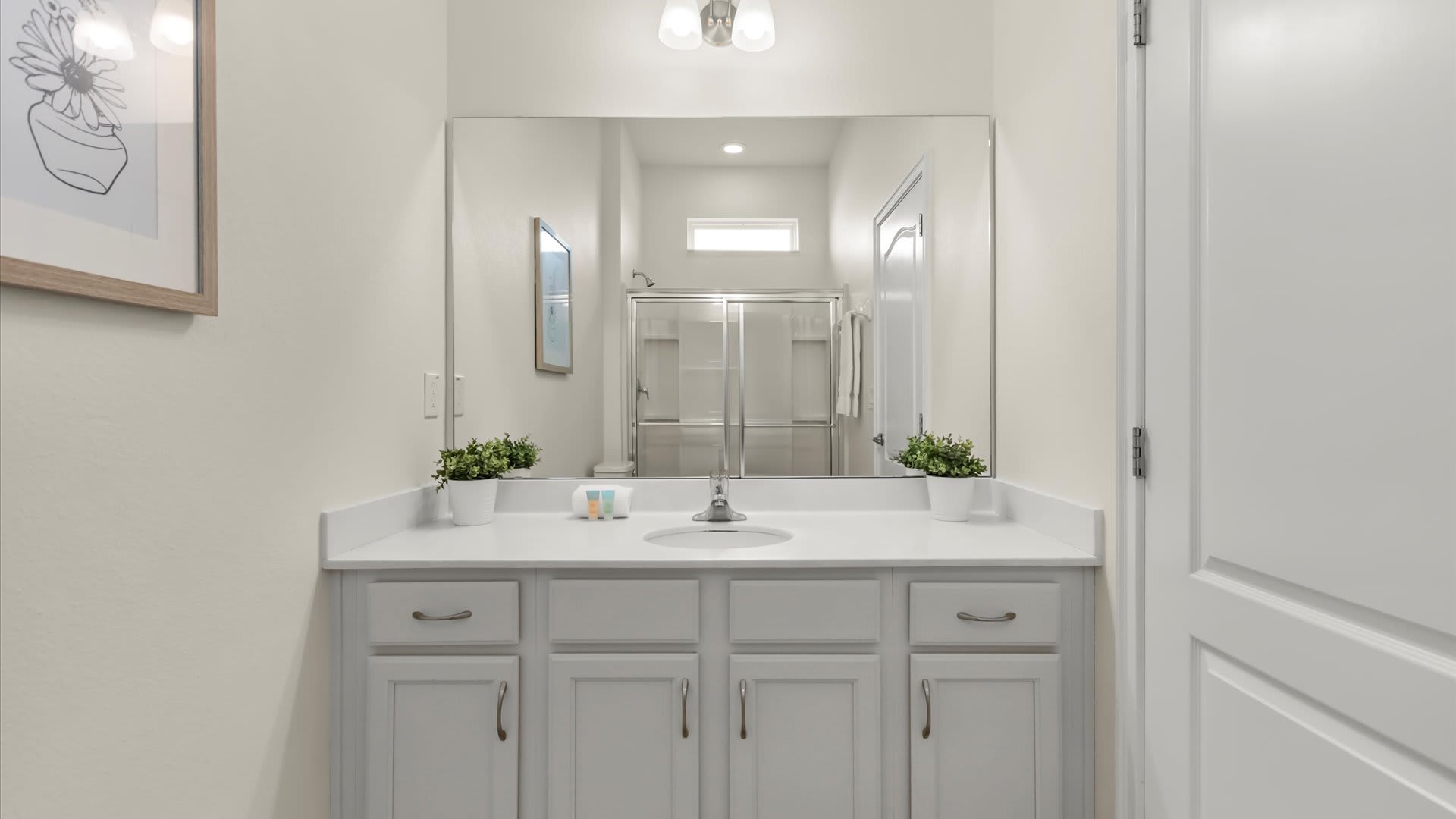 A bright, modern bathroom featuring a single vanity with a white countertop, light gray cabinets, and a large mirror reflecting a glass-enclosed shower. Two small potted plants add a touch of greenery.