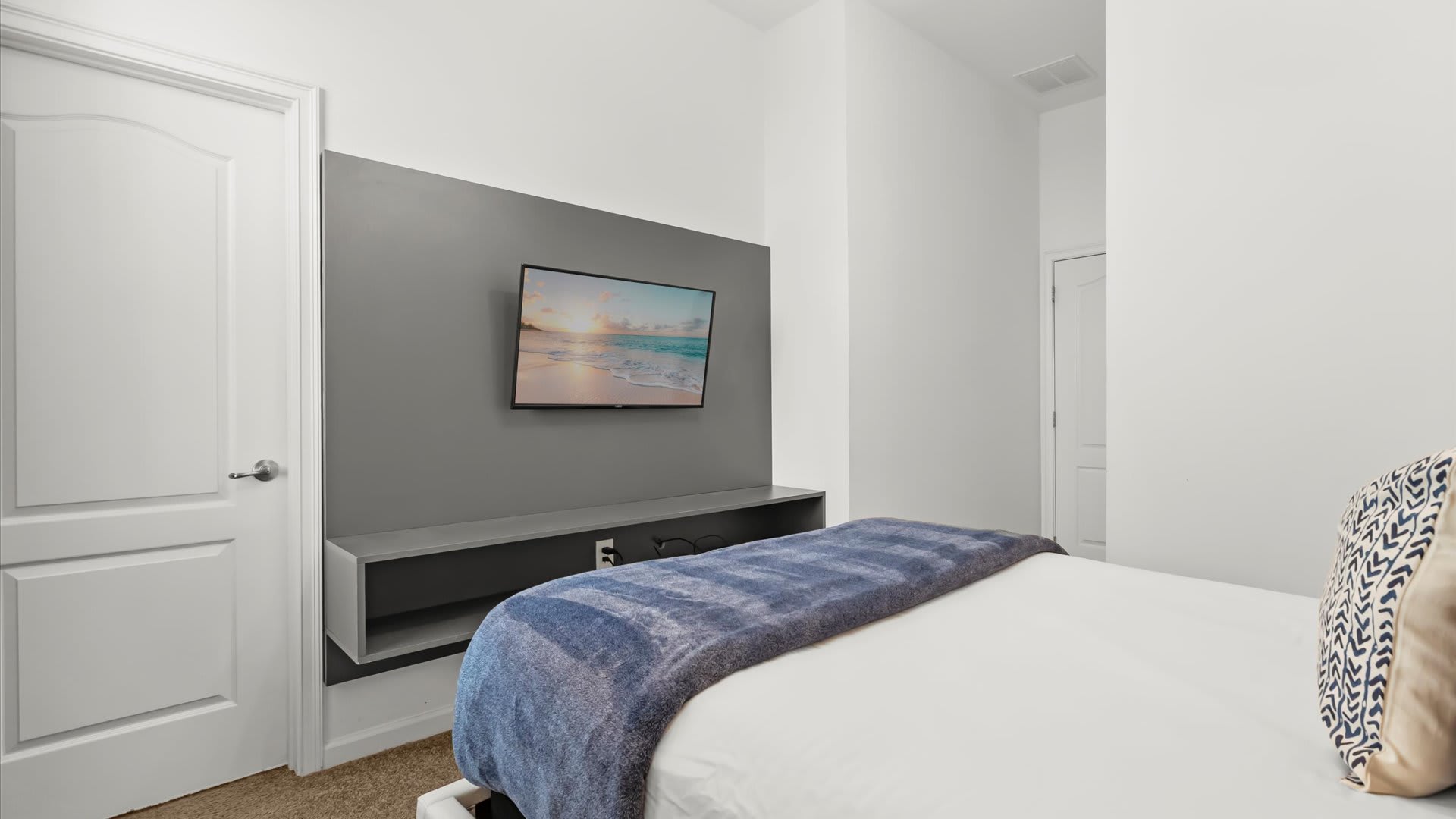 A modern bedroom wall featuring a flat-screen TV mounted on a grey accent panel with a floating shelf below, positioned opposite a bed with a blue blanket. A white door is visible to the left.