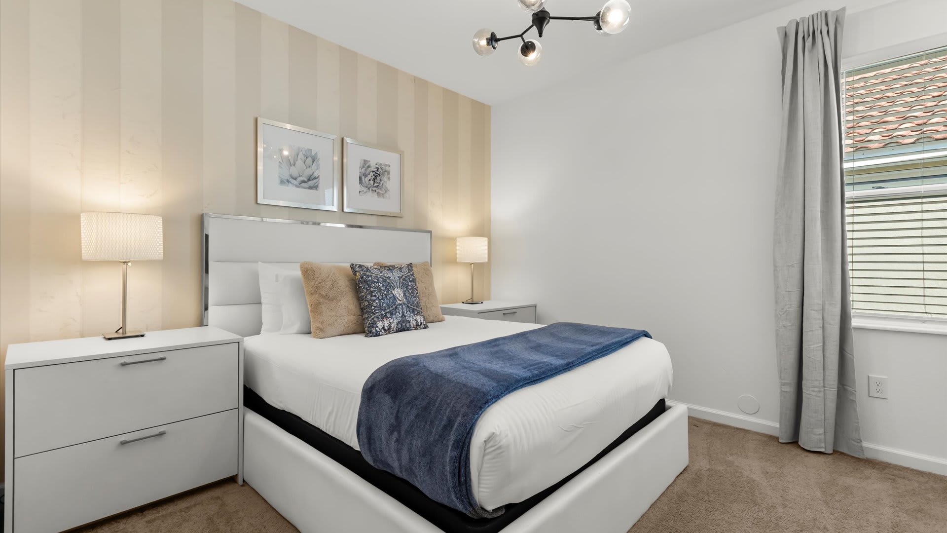 A stylish bedroom featuring a queen-sized bed with a white upholstered headboard, two white nightstands with lamps, and a striking accent wall with vertical beige and cream stripes. A modern chandelier hangs from the ceiling.