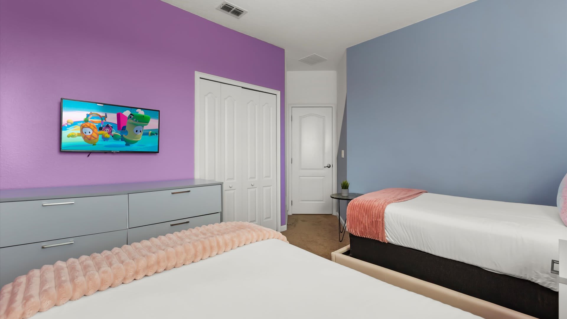 View of the themed bedroom with two twin beds, showing a wall-mounted TV displaying a cartoon, a white bi-fold closet, and a purple accent wall with a gray dresser.