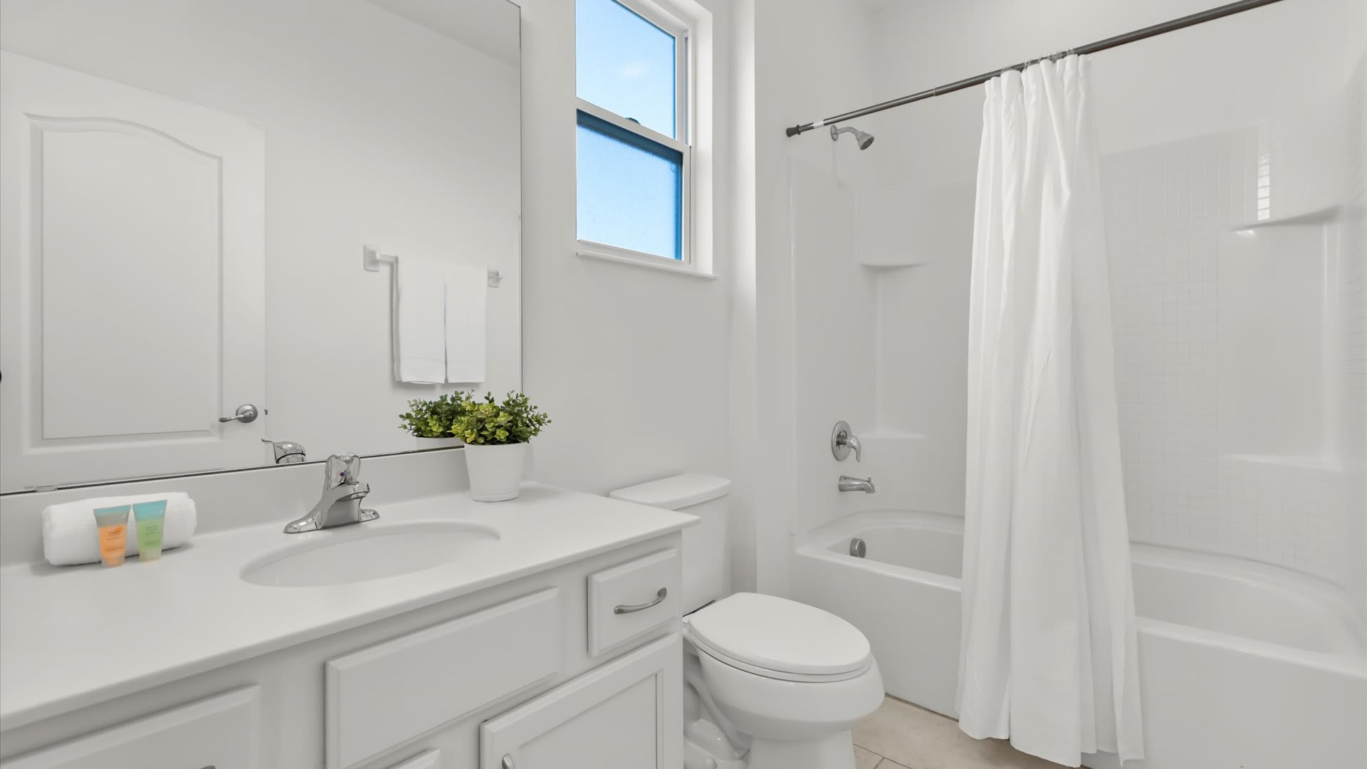 A well-lit bathroom with a single vanity, white countertop, and a toilet. A shower-tub combination with a white curtain is visible, and a small window provides natural light.