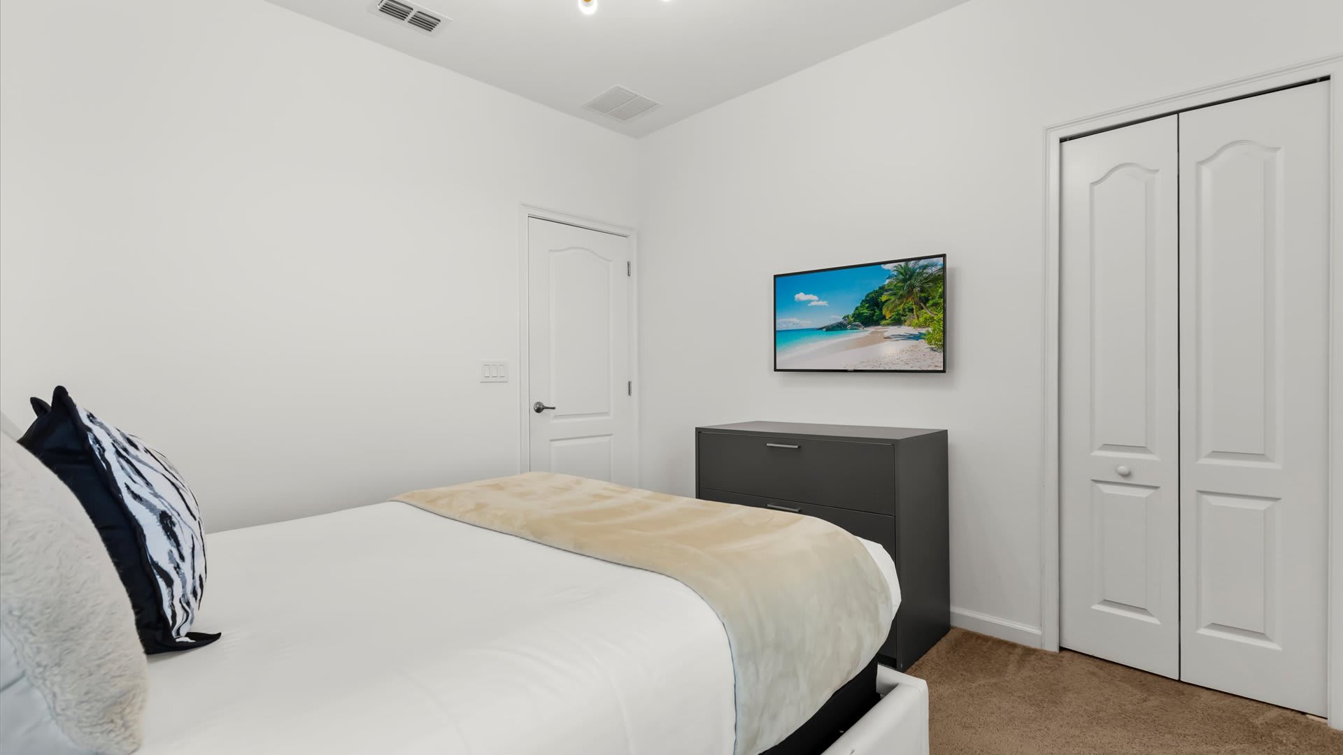 View of the foot of the bed in bedroom-2, showing a dark gray dresser with a wall-mounted flat-screen TV displaying a beach scene. A white bi-fold closet door is visible to the right.