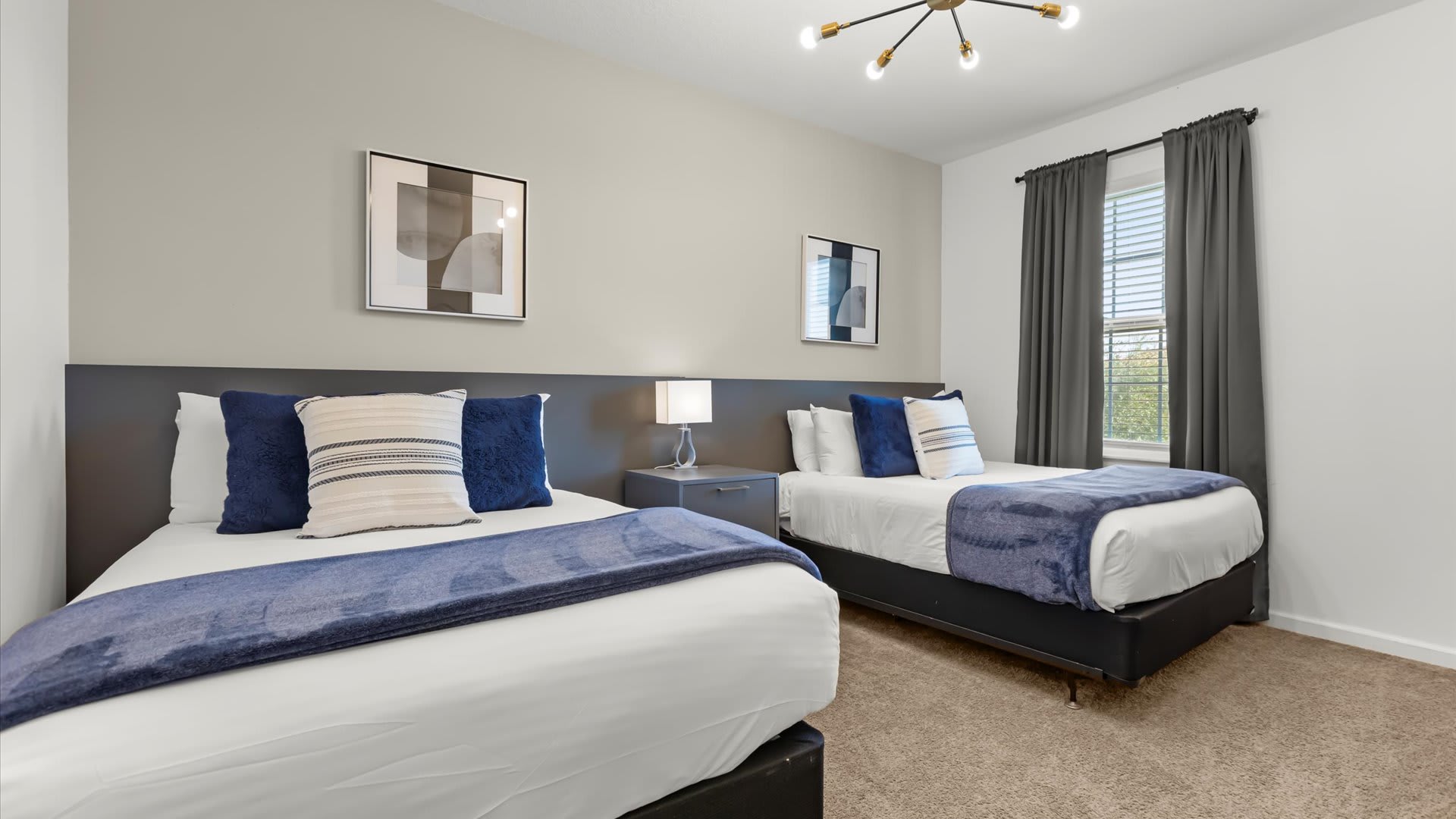 Serene bedroom with two twin beds, each featuring blue throw blankets and pillows, set against a light gray wall with modern framed artwork. Dark curtains frame a window.