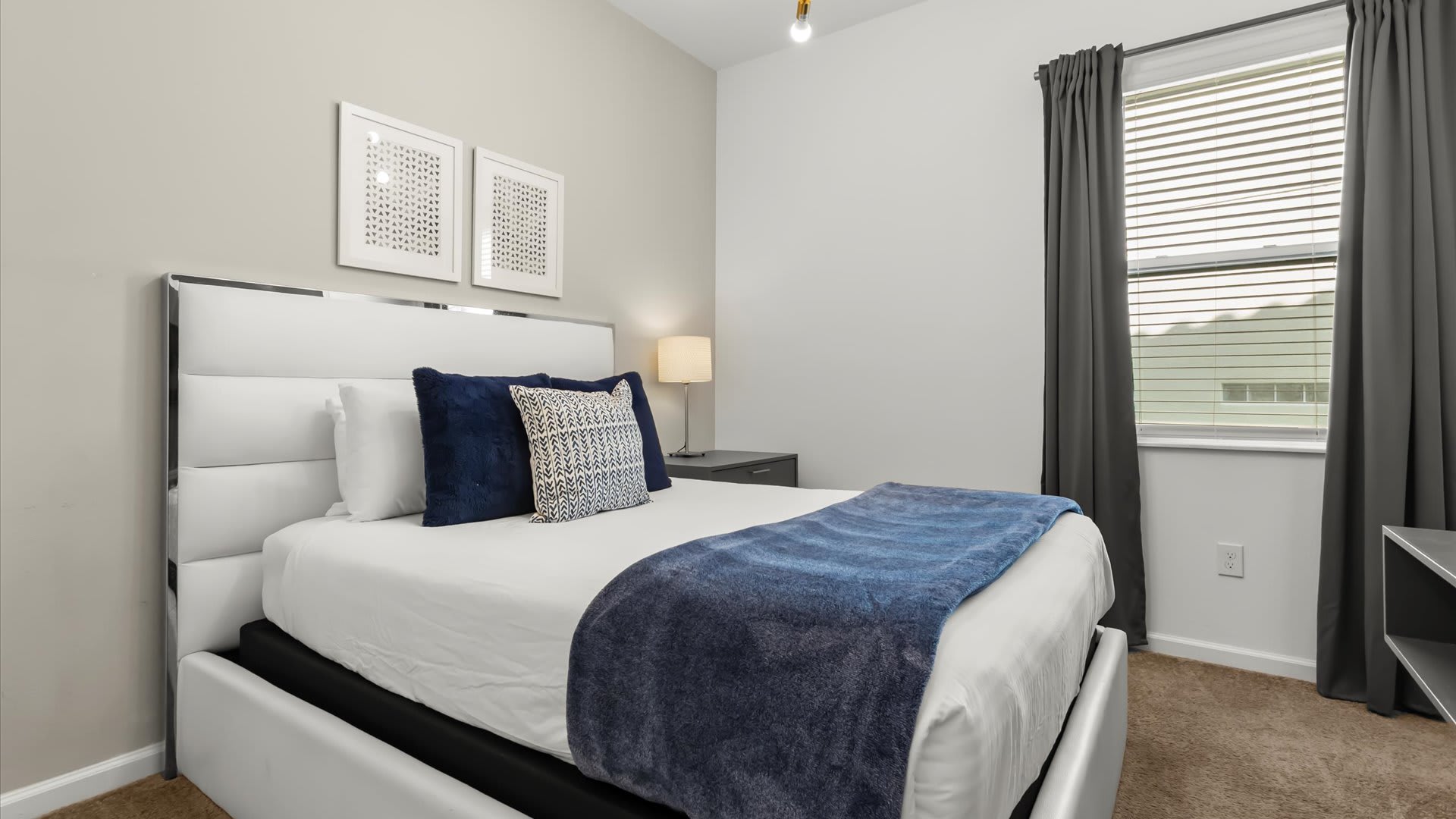 A cozy bedroom featuring a white upholstered queen bed with a blue throw blanket and decorative pillows, flanked by a dark grey nightstand with a lamp. The room has light grey walls and a window with dark grey curtains.