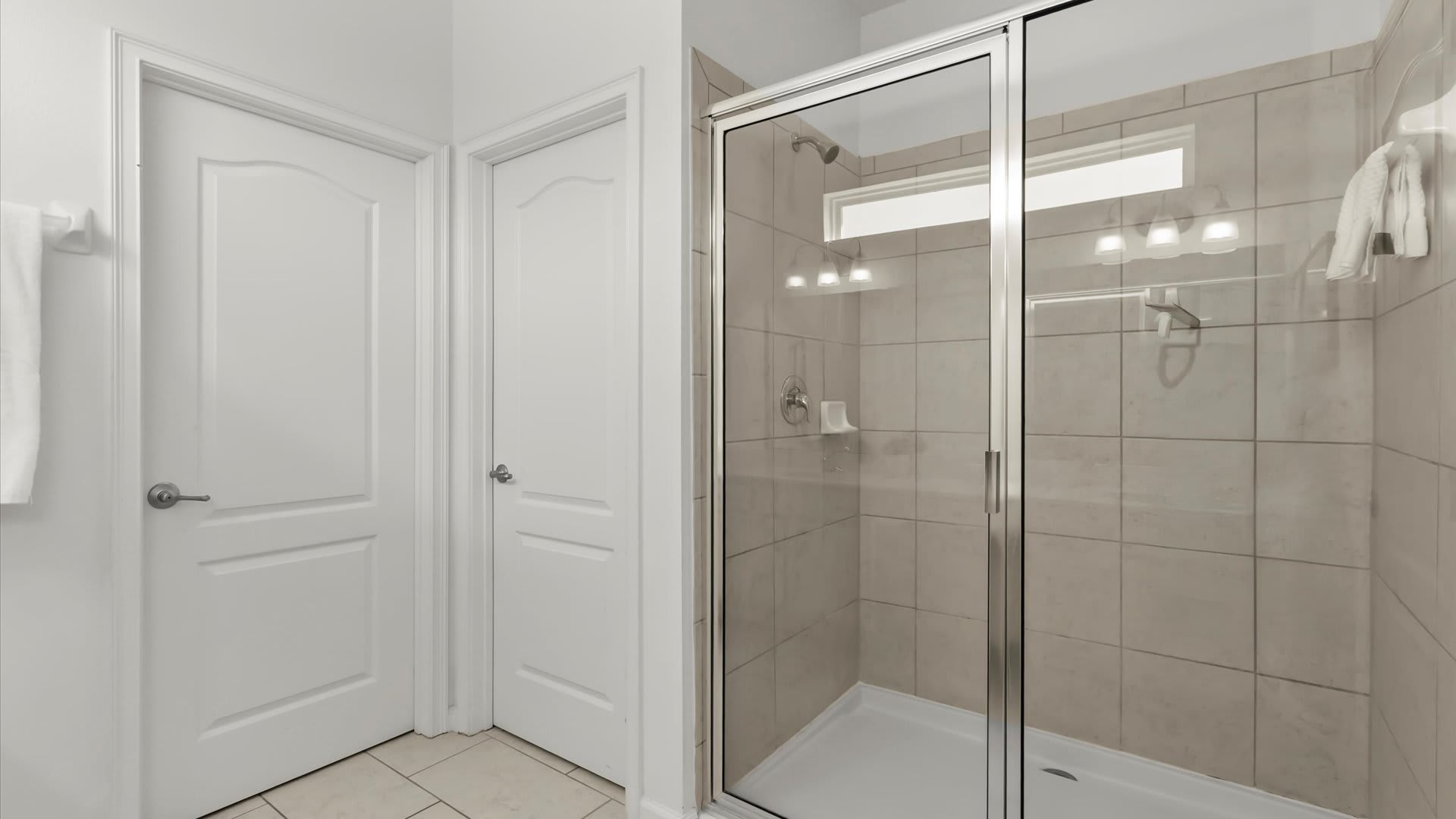 Close-up view of the walk-in shower in bathroom-1, featuring light beige subway tiles, a glass enclosure, and a small horizontal window above. Two white doors are visible to the left.