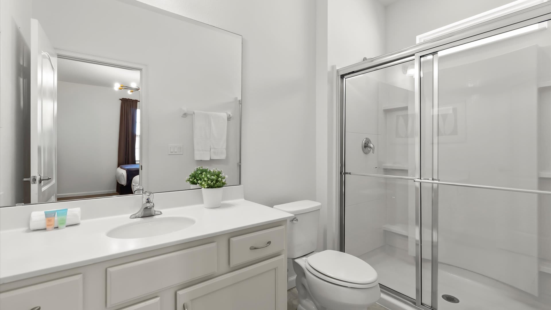 Modern white bathroom featuring a single vanity with a large mirror, a toilet, and a spacious glass-enclosed shower. The mirror reflects a glimpse of a bedroom with blue accents.
