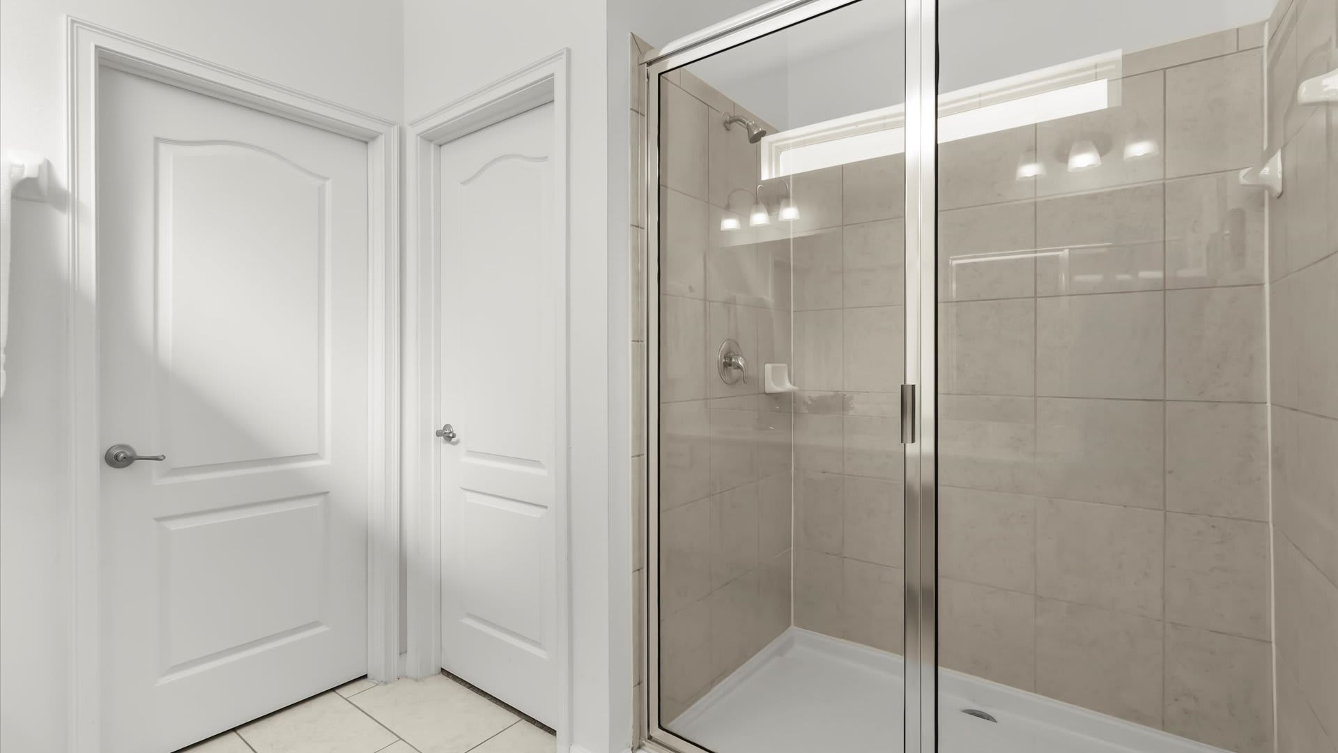 A modern walk-in shower with clear glass doors and light-colored square tiles covering the walls, featuring a chrome showerhead and fixtures. The shower base is white, and the floor outside is tiled.