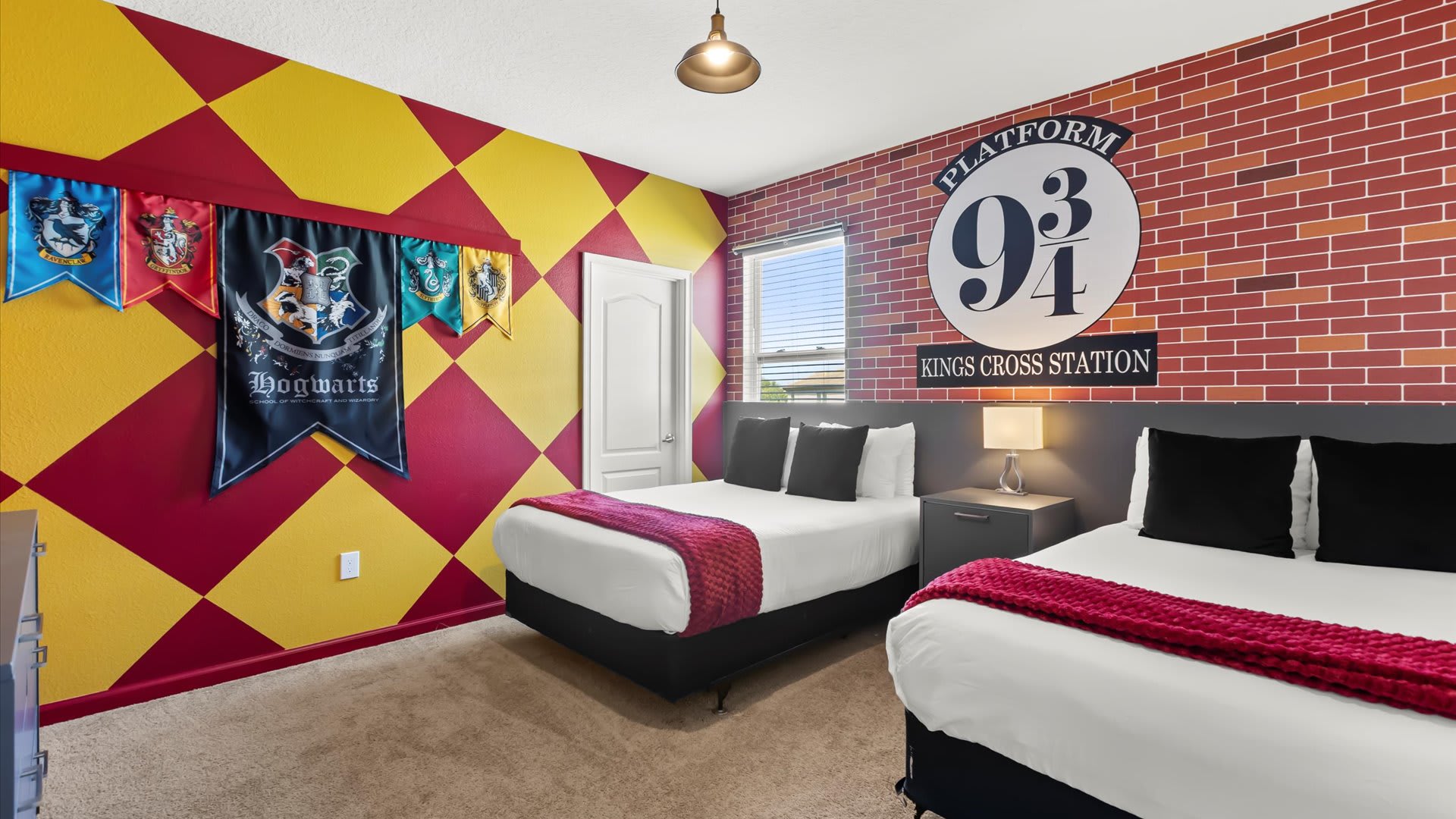 A vibrant Harry Potter-themed bedroom featuring two queen beds, a red and yellow diamond-patterned accent wall, and a "Platform 9 3/4 Kings Cross Station" sign above one bed. Hogwarts house banners hang on the wall.