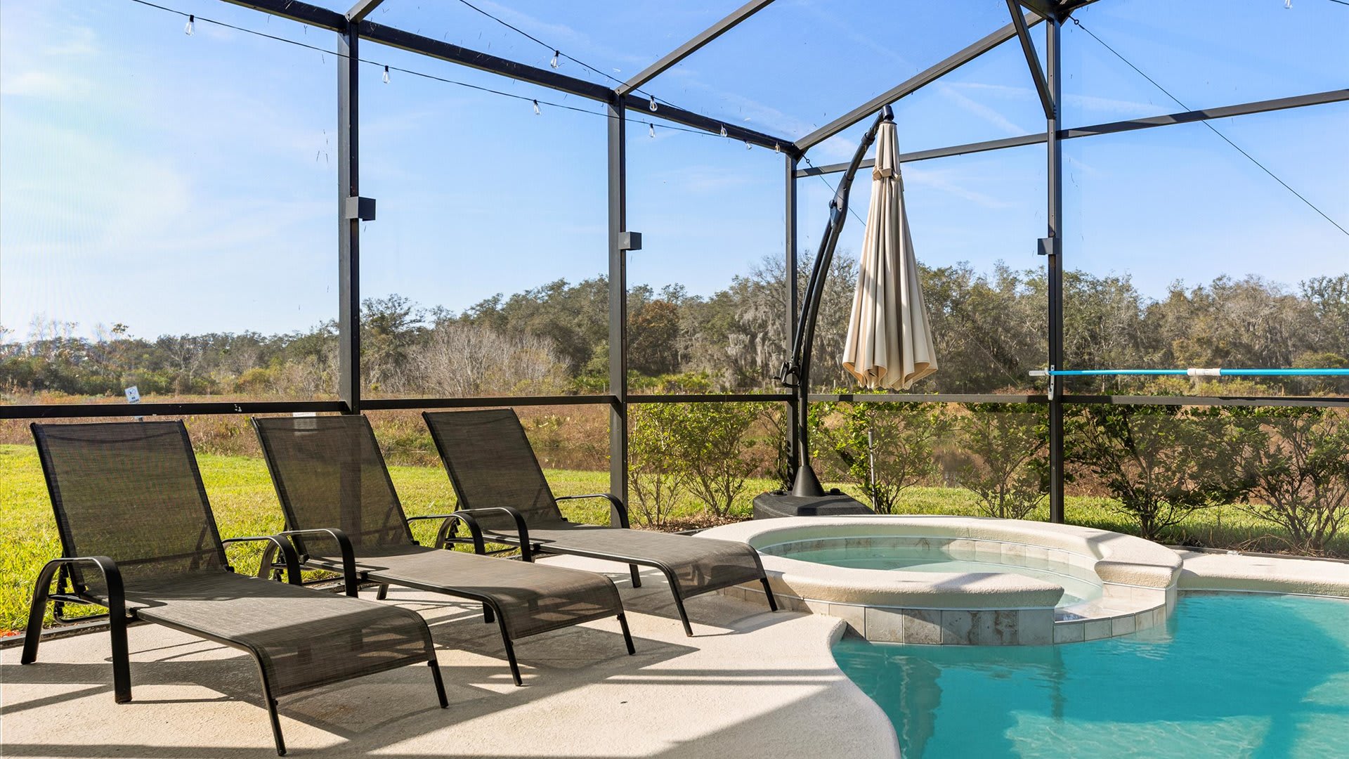 Sunny view of the private screened pool and hot tub, featuring several dark mesh lounge chairs and a large patio umbrella, with a serene natural landscape visible beyond the enclosure.