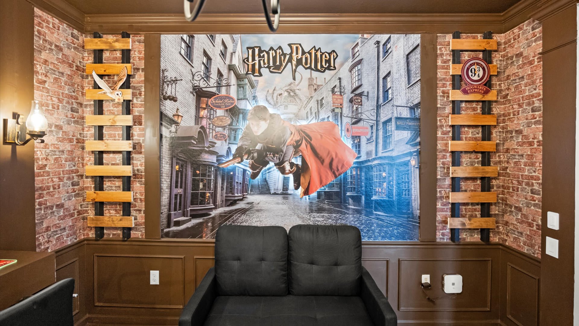 A Harry Potter-themed game room features a large mural of Harry Potter flying over Diagon Alley, flanked by decorative wooden ladders and brick-patterned walls. A black sofa provides seating in front of the mural, with dark wood wainscoting completing the themed decor.