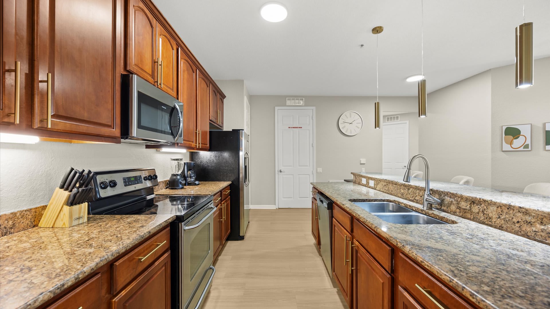 A galley-style kitchen showcases dark wood cabinetry, speckled granite countertops, and stainless steel appliances, including a double sink and dishwasher. The light wood flooring extends throughout, with a wall clock visible in the background and a breakfast bar on the right.