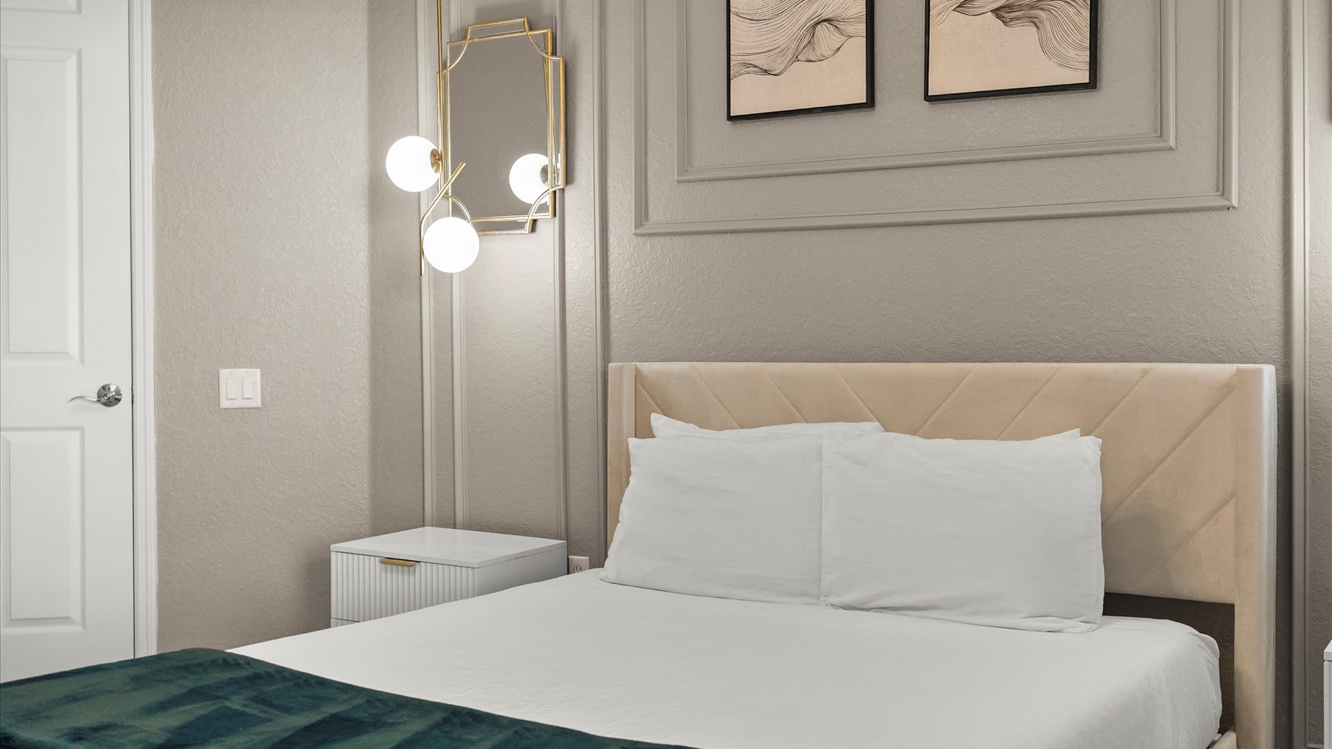 A modern bedroom features a bed with a light beige upholstered headboard and white bedding, accented by a dark green throw blanket. A white nightstand with a ribbed drawer sits beside the bed, illuminated by a gold geometric pendant light, against a light gray wall with decorative molding.