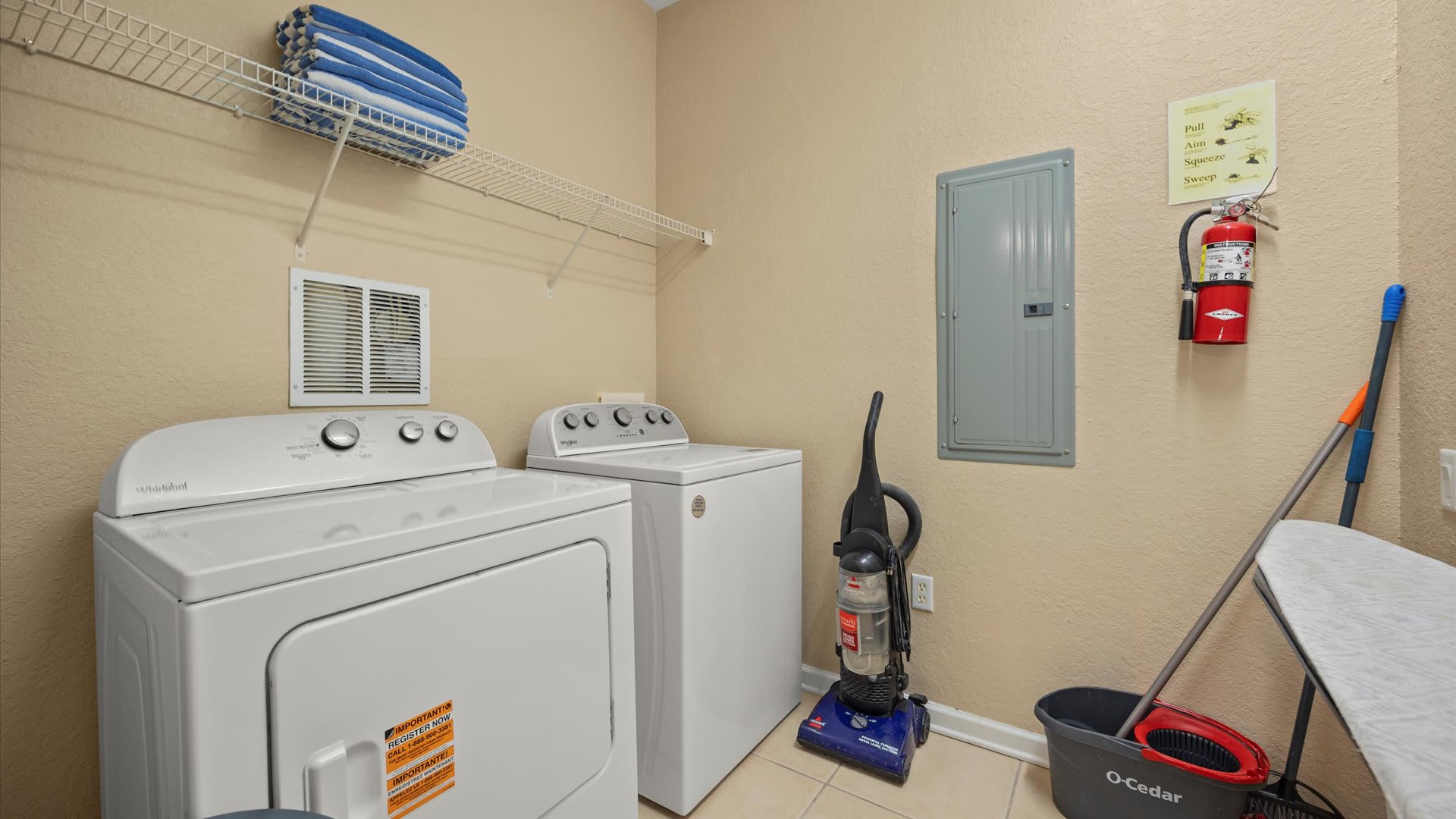 A dedicated laundry closet features a white top-loading washing machine and a front-loading dryer, with a wire shelf holding folded blue towels above. A vacuum cleaner, cleaning supplies, and a fire extinguisher are also present.