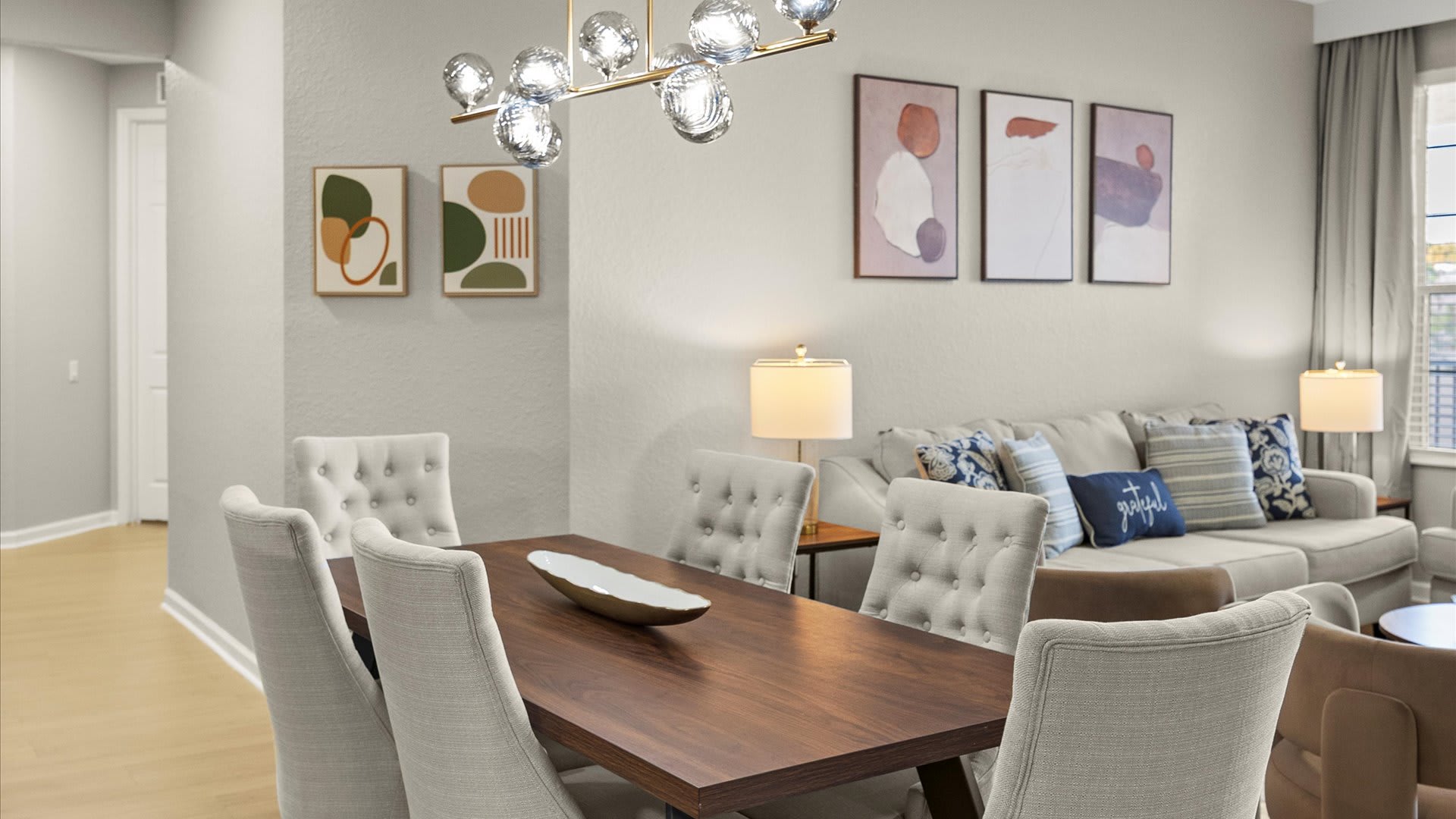 A modern dining area features a dark wood rectangular table surrounded by six light-colored upholstered chairs with tufted backs, illuminated by a contemporary glass orb chandelier. In the background, a living room with a light gray sofa and decorative pillows is visible, along with abstract wall art.