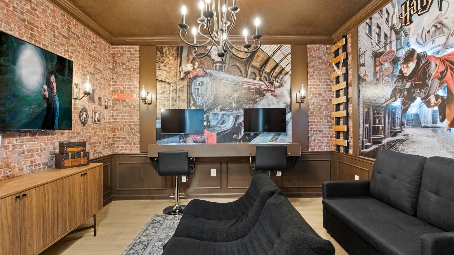 A Harry Potter-themed game room features brick-patterned wallpaper, dark wood wainscoting, and a large mural of the Hogwarts Express. Two black gaming chairs are positioned in front of dual monitors, with two black sofas providing additional seating, all under a dark chandelier.