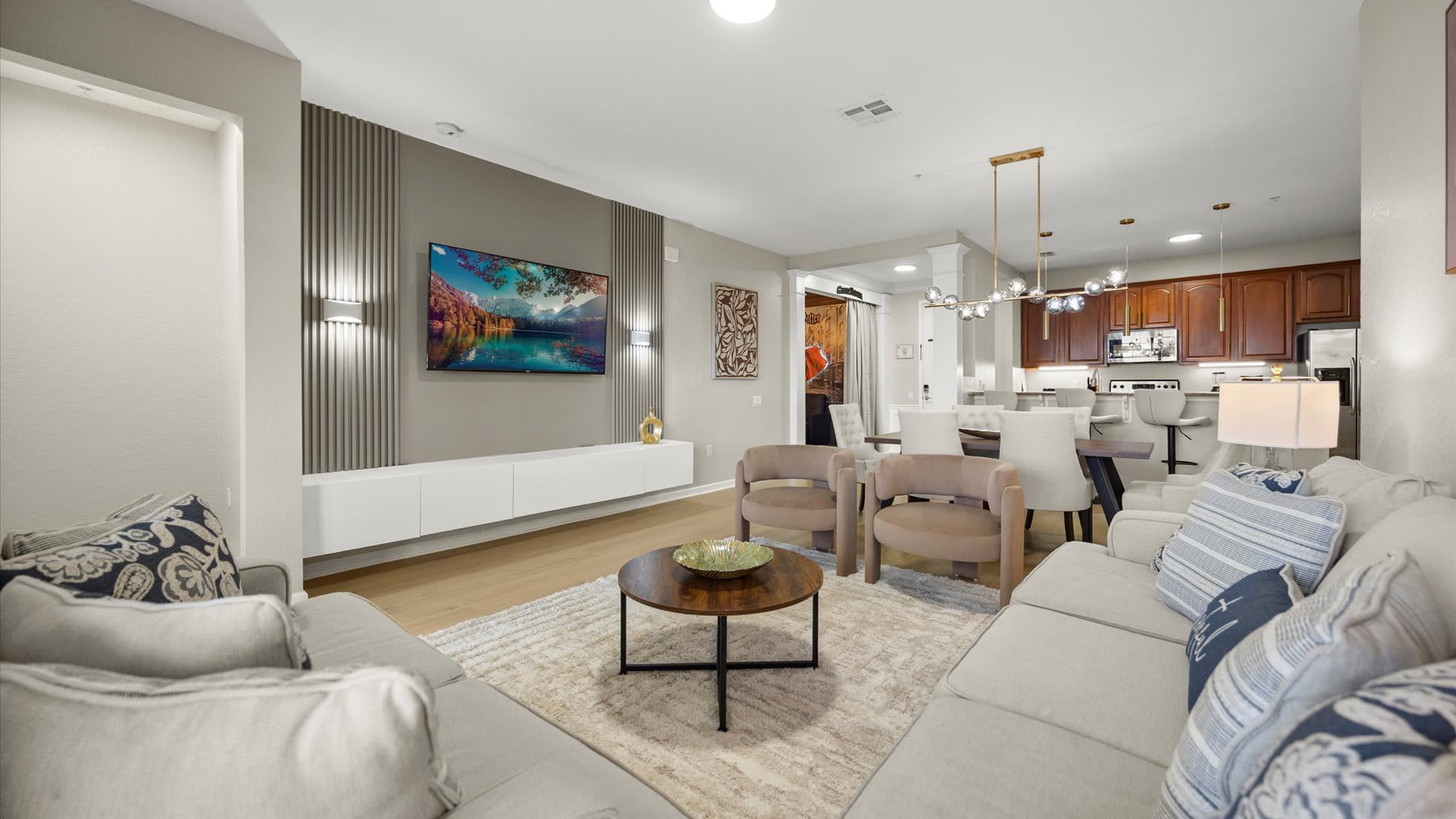 An expansive open-concept living space seamlessly connecting the living room, dining area, and kitchen. The living room features light-colored sofas and armchairs facing a TV mounted on a grey accent wall, while the dining area and kitchen with dark wood cabinets are visible in the background.
