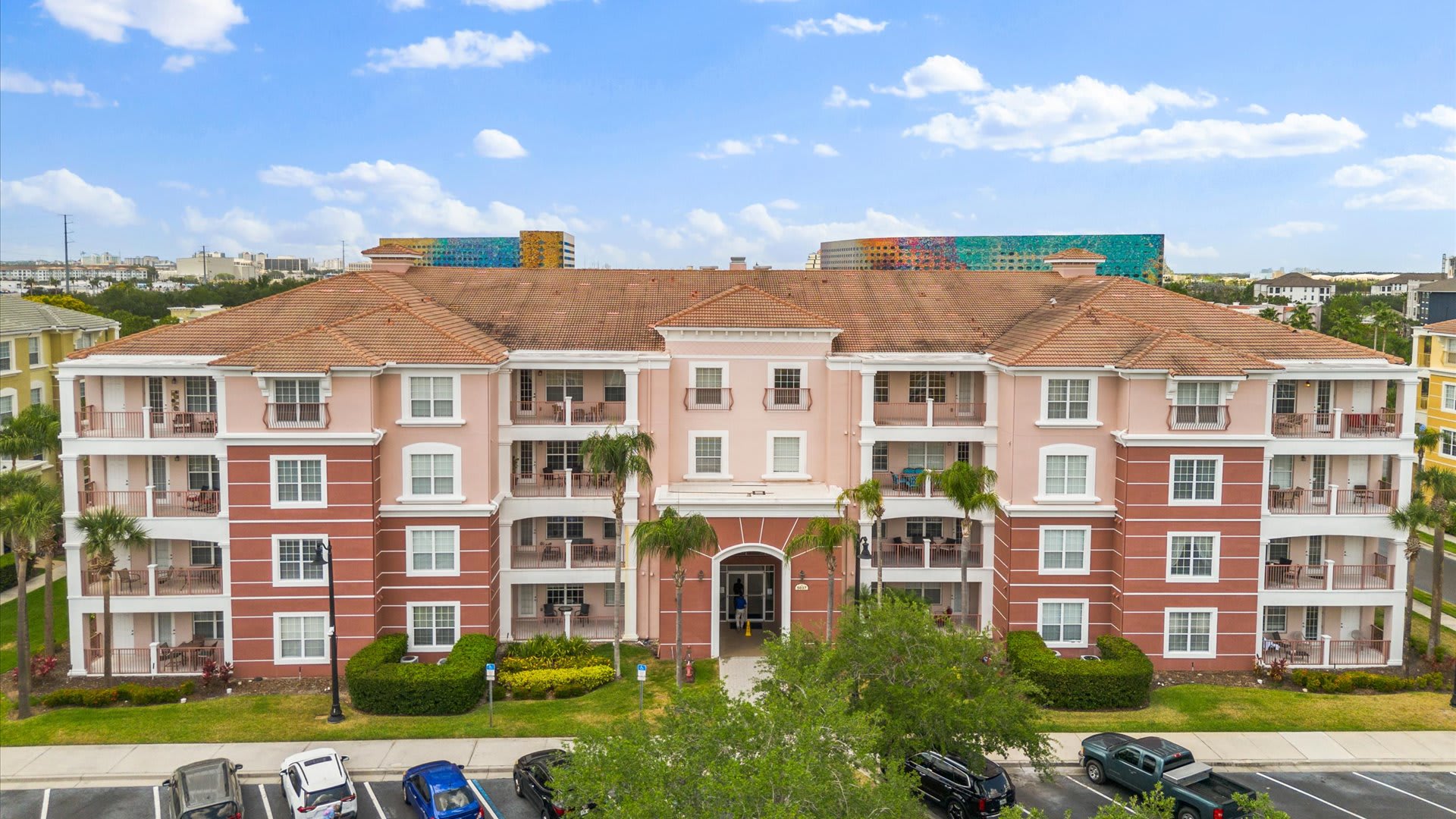 A large, multi-story condominium building with a pink and brick-red facade, featuring numerous balconies and a classic tiled roof. Palm trees line the front, and a parking lot is visible in the foreground.