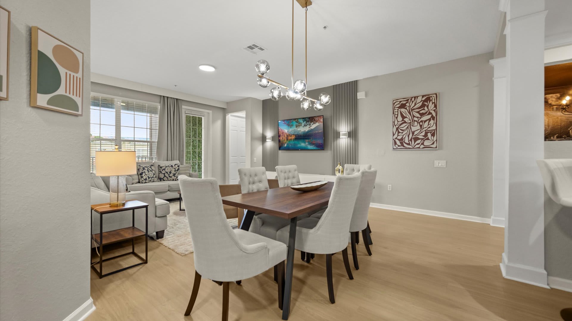 A modern dining area with a dark wood table and six upholstered chairs, illuminated by a linear chandelier, positioned in front of a grey accent wall with a mounted TV. The space opens to a living room on the left and a kitchen with dark wood cabinetry on the right.