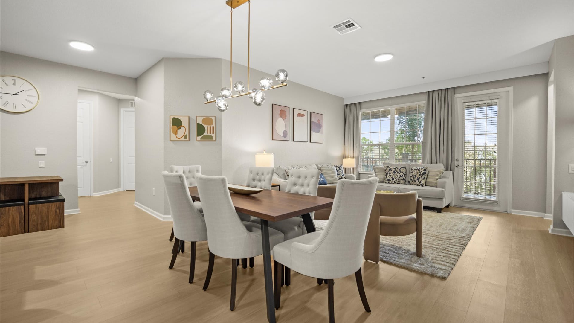 An open-concept dining area featuring a dark wood dining table with six light-colored upholstered chairs, illuminated by a modern linear chandelier. The space flows into a living room with sofas and armchairs, all set on light wood flooring.