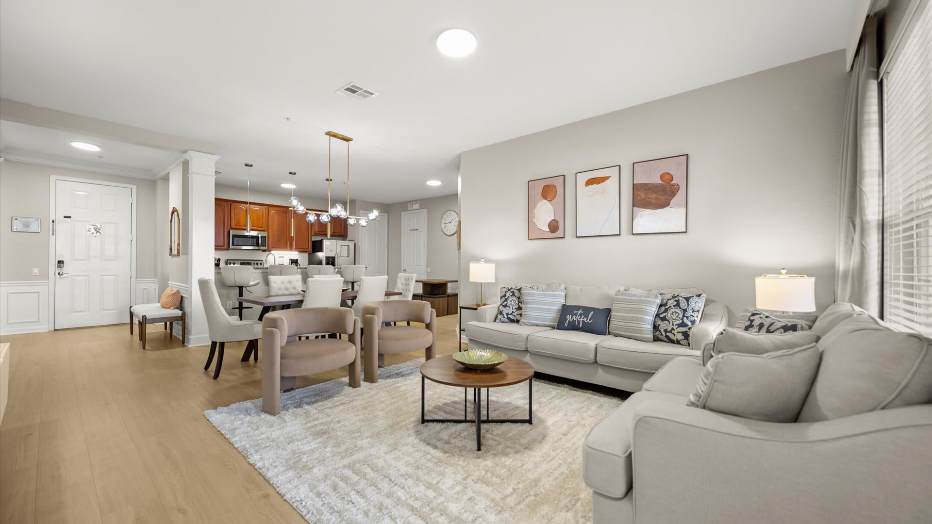 A spacious open-concept area showcasing a living room with light-colored sofas and armchairs, flowing into a dining space with a dark wood table and upholstered chairs under a modern chandelier. The entrance door and a wall clock are visible in the background, all on light wood flooring.