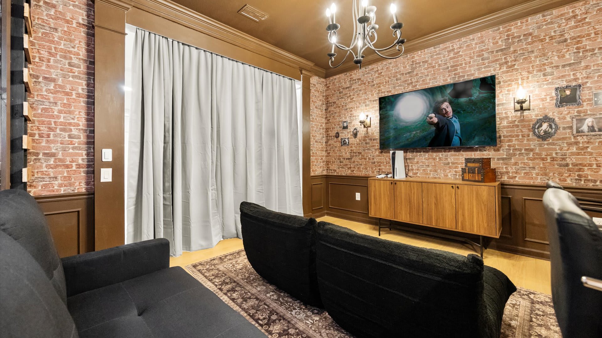 A Harry Potter-themed game room features brick-patterned walls, dark wood wainscoting, and a large flat-screen TV displaying a movie. Two black sofas are arranged on an area rug, with a wooden media console below the TV and a chandelier providing overhead lighting.