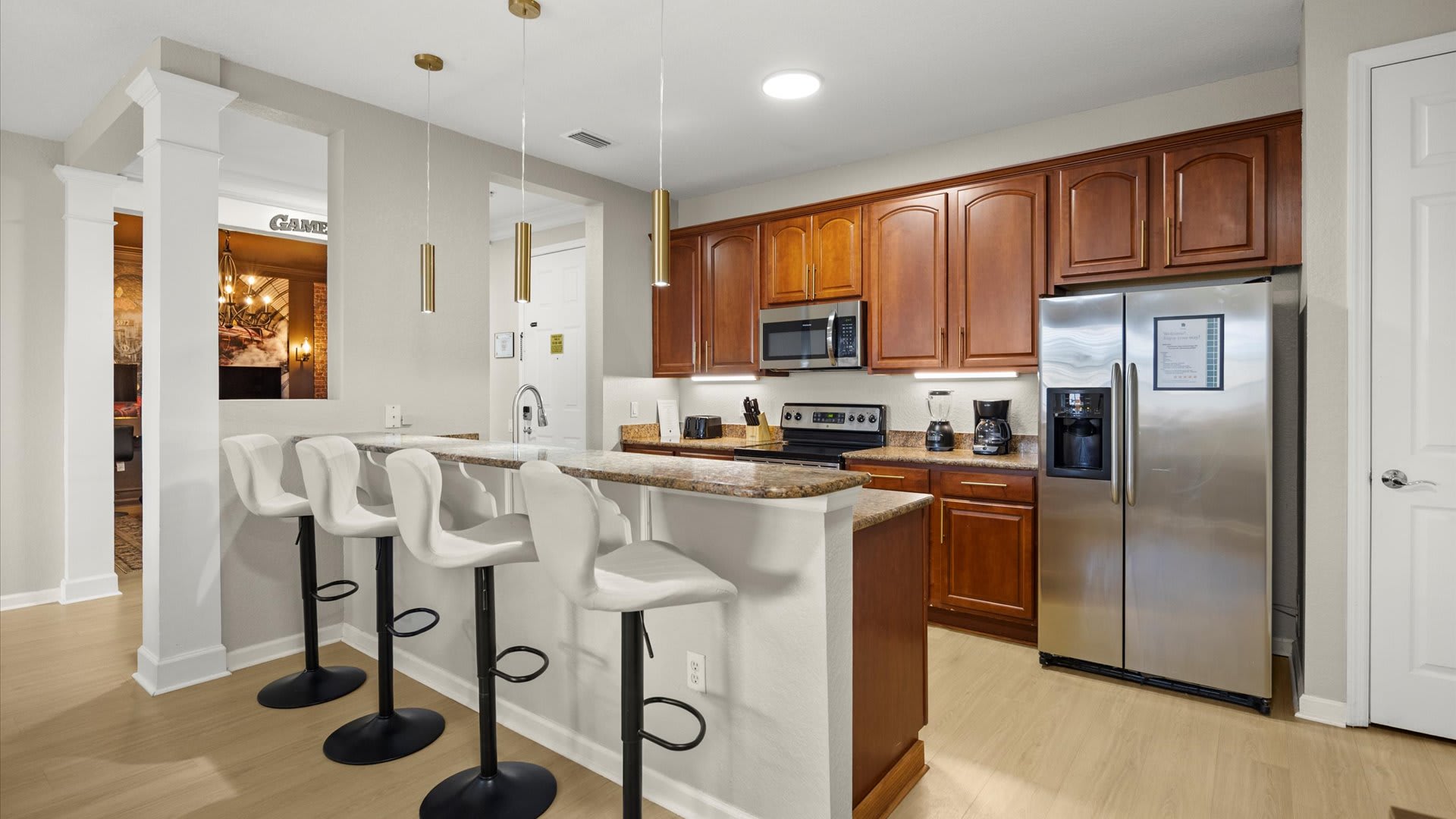 An open-concept kitchen features dark wood cabinets, granite countertops, and stainless steel appliances, including a refrigerator, microwave, and stove. A breakfast bar with four white and black bar stools is highlighted by three gold pendant lights, connecting the kitchen to an adjacent living area.